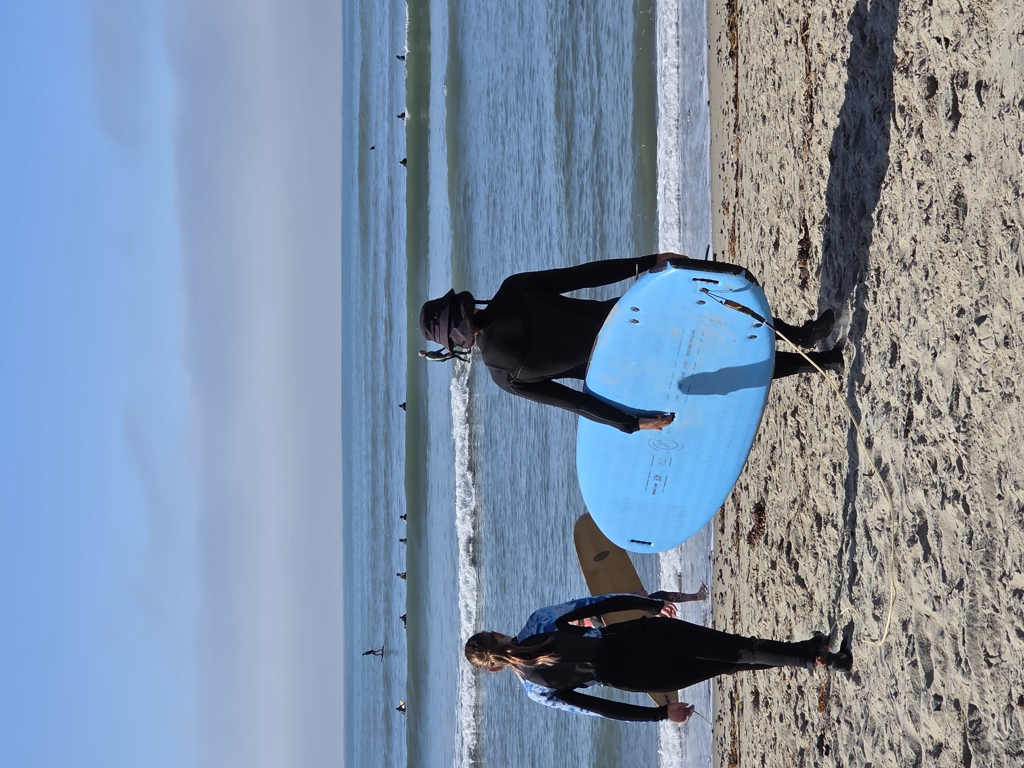 Two individuals in wetsuits are walking on a sandy beach, carrying surfboards towards the ocean in Dana Point, United States of America. The person on the left, facing away from the camera, wears a blue and black wetsuit and carries a light brown surfboard. The person on the right, also facing away, is dressed in a black wetsuit and a dark hat, carrying a bright blue surfboard. Several other surfers are visible in the ocean, some riding waves and others waiting for them. The water is blue with white-capped waves, and the sky is clear and blue with some distant clouds. The beach is covered in sand and some seaweed.