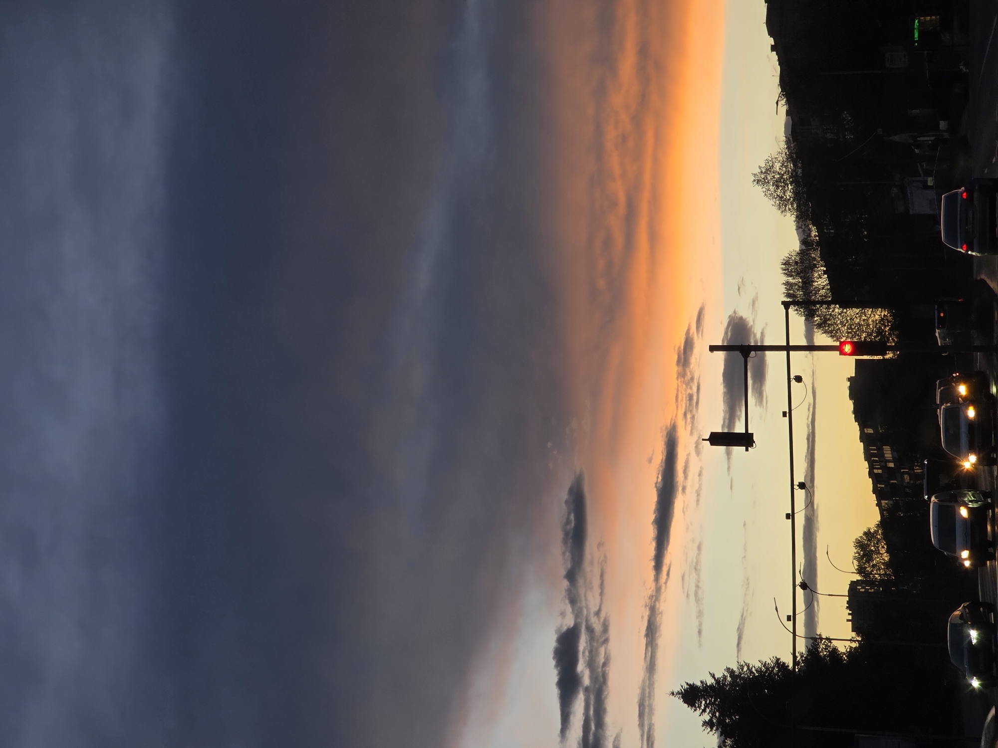 The image captures a street scene at twilight in Varna, Bulgaria. The sky is divided into two distinct regions: the left side is dominated by dark, overcast clouds, while the right side displays a vibrant sunset with orange and yellow hues. A traffic light is visible in the center of the frame, displaying a red light. Cars with headlights on are stopped at the intersection. Silhouetted buildings and trees line the horizon on the right side of the image. The street appears to be relatively busy with traffic.
