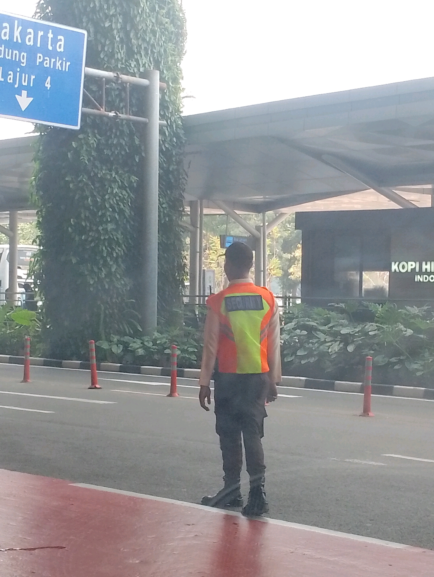 A security guard stands on a paved road in front of a building with "KOPI HI" and "INDO" visible on a dark facade. The guard wears a reflective vest with "SECURITY" printed on the back in a yellow rectangle and dark pants and boots. To the left of the guard, a blue directional sign indicates "Jakarta", "Bandung Parkir", and "Lajur 4" with a downward arrow. Orange traffic cones are spaced along the road's edge. Lush green foliage covers a large pillar behind the sign. The scene appears to be outdoors during daylight.