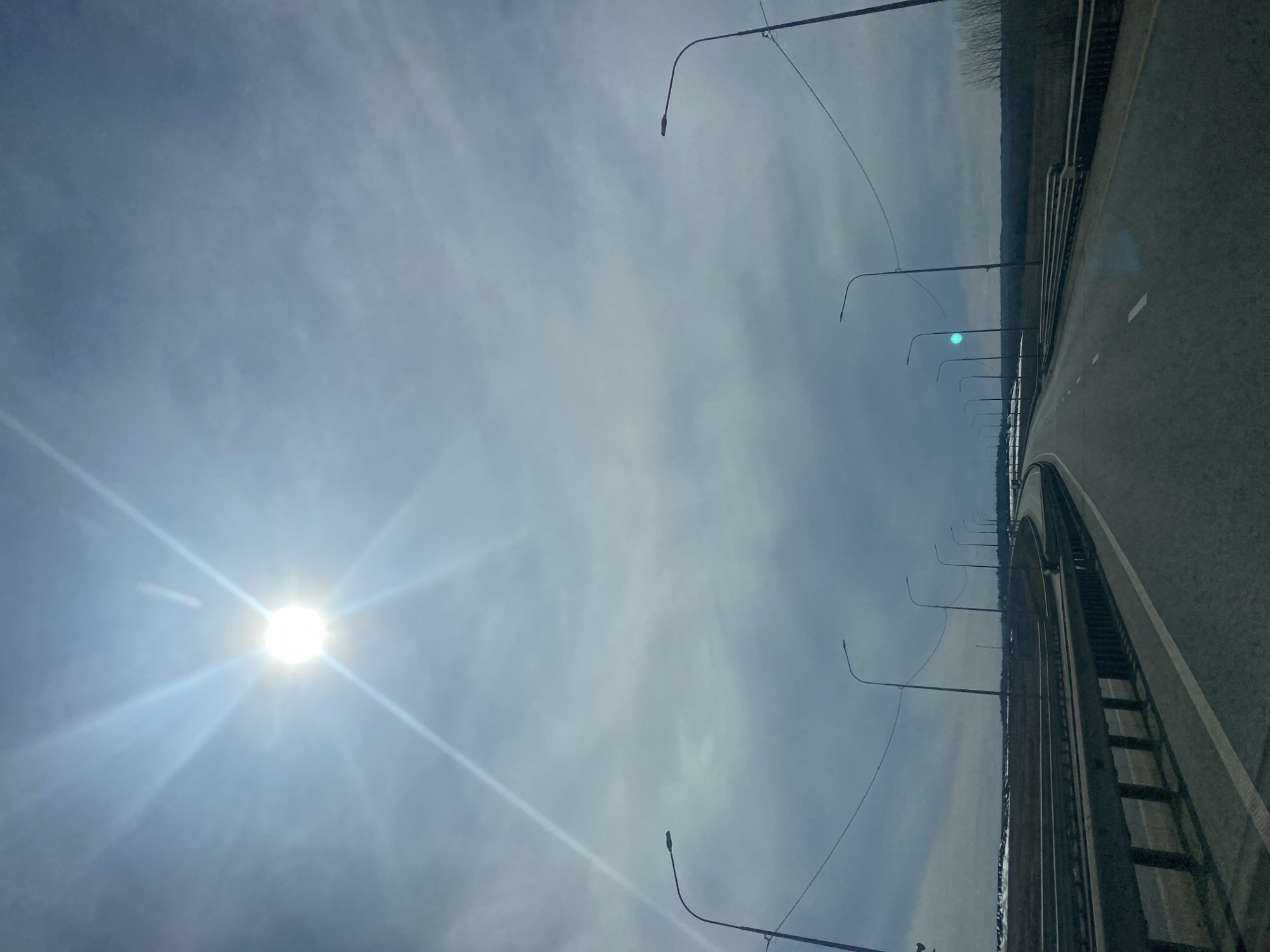 The image depicts a section of roadway in Kliuchiki, Russia. The road is gray and features white lane markings. A metal guardrail is present along the right edge of the roadway. A row of streetlights lines the left side. Beyond the guardrail on the right is a wooded area. The sky is overcast with a bright sun visible on the left.