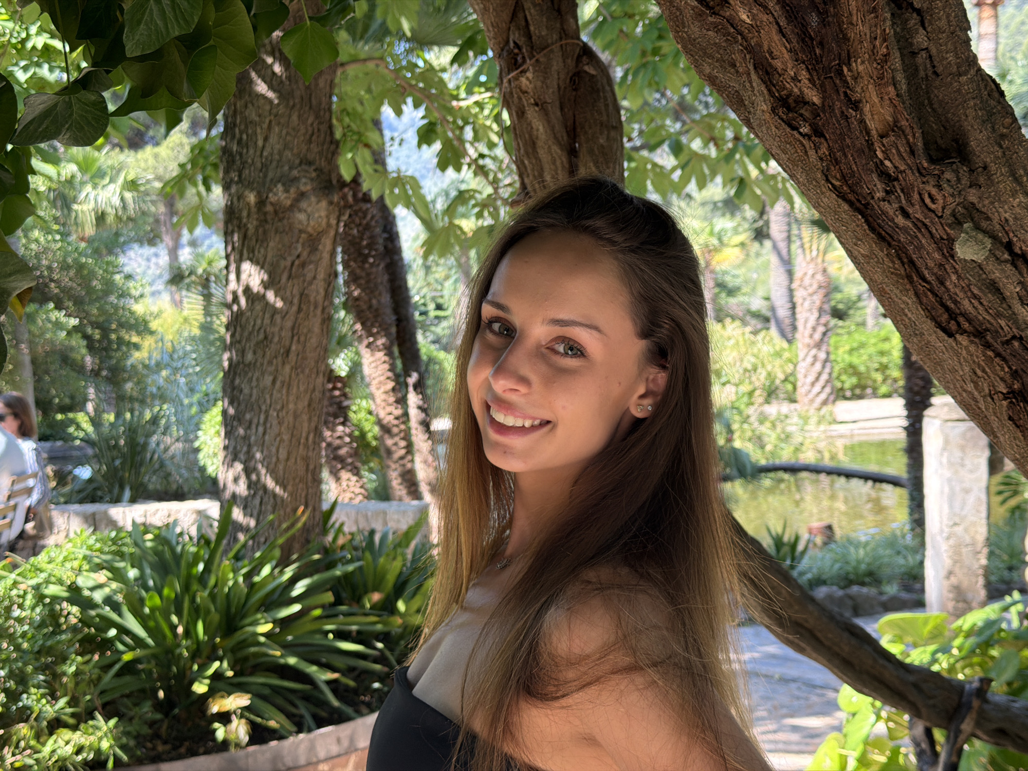 The photograph depicts a young woman with long brown hair and fair skin, smiling directly at the camera. She is positioned in what appears to be a park or garden, framed by tree trunks and foliage. In the background, glimpses of a pond or body of water can be seen, along with other trees and vegetation, suggesting a lush, natural environment. A partial view of people sitting on a bench is visible at the left side of the image. Bunyola, Spain is not directly identifiable within the photo; however, the park's design and landscape elements align with Mediterranean park or garden features.