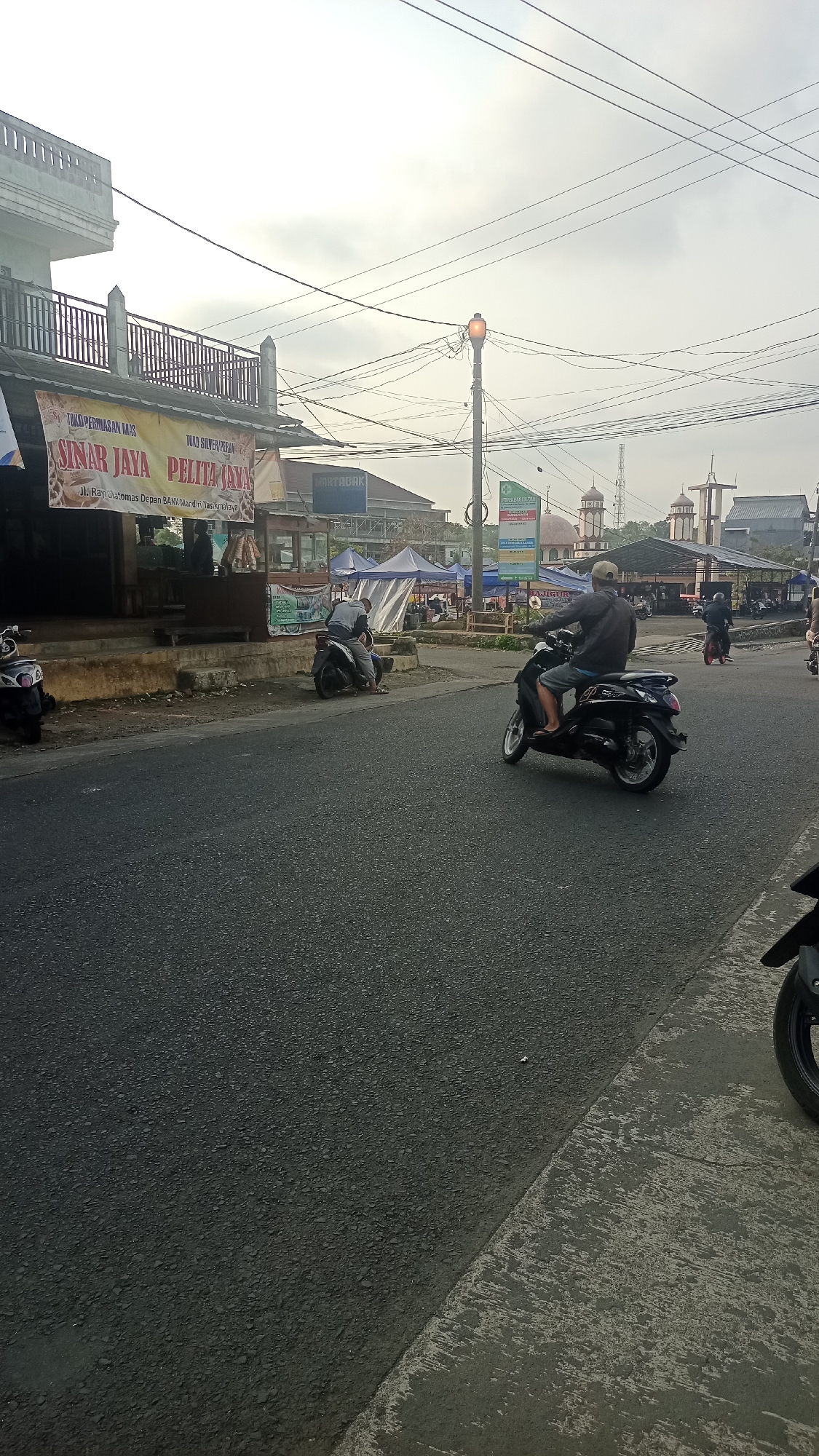 The image depicts a street scene in Karyamandala, Indonesia, with a cloudy sky. In the foreground, a dark asphalt road occupies the majority of the frame, bordered by a textured concrete shoulder on the right. Several motorcycles are visible: one is parked on the left side, another is being ridden by a person wearing a grey jacket and shorts in the middle of the road, and a third, red motorcycle with a rider is further down the street on the right.
Along the left side of the road, there is a building with a white upper floor and a lower level housing shops. A prominent sign above the shops reads "SINAR JAYA PELITA JAYA" with additional text indicating "JL. Raya Ciomas Depan BANK MANDIRI Tasikmalaya". Tents are set up in front of some of the shops, suggesting a market or roadside stalls.
Further in the background, more buildings and structures are visible, including what appear to be mosque domes, indicating a community with religious diversity. Power lines crisscross the sky, supported by poles, including one with an illuminated orange traffic signal. The overall impression is that of a busy, everyday street in a populated area of Indonesia.