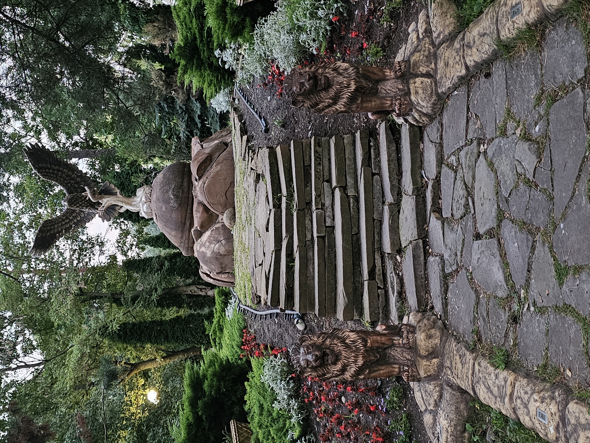 The image depicts an ornate garden pathway leading up a stone staircase, flanked by two lion statues. At the top of the stairs, an eagle sculpture stands majestically atop a rocky base, with its wings spread wide. The garden is lush with greenery, including manicured bushes, flowering plants, and tall trees. The pathway is made of irregular stone slabs, and the steps are also constructed from large, flat stones. The overall impression is one of a carefully designed and artfully arranged landscape. This scene is located at the "Cherkessk City Park" in Cherkessk, Russia, a public green space known for its sculptures and natural beauty.