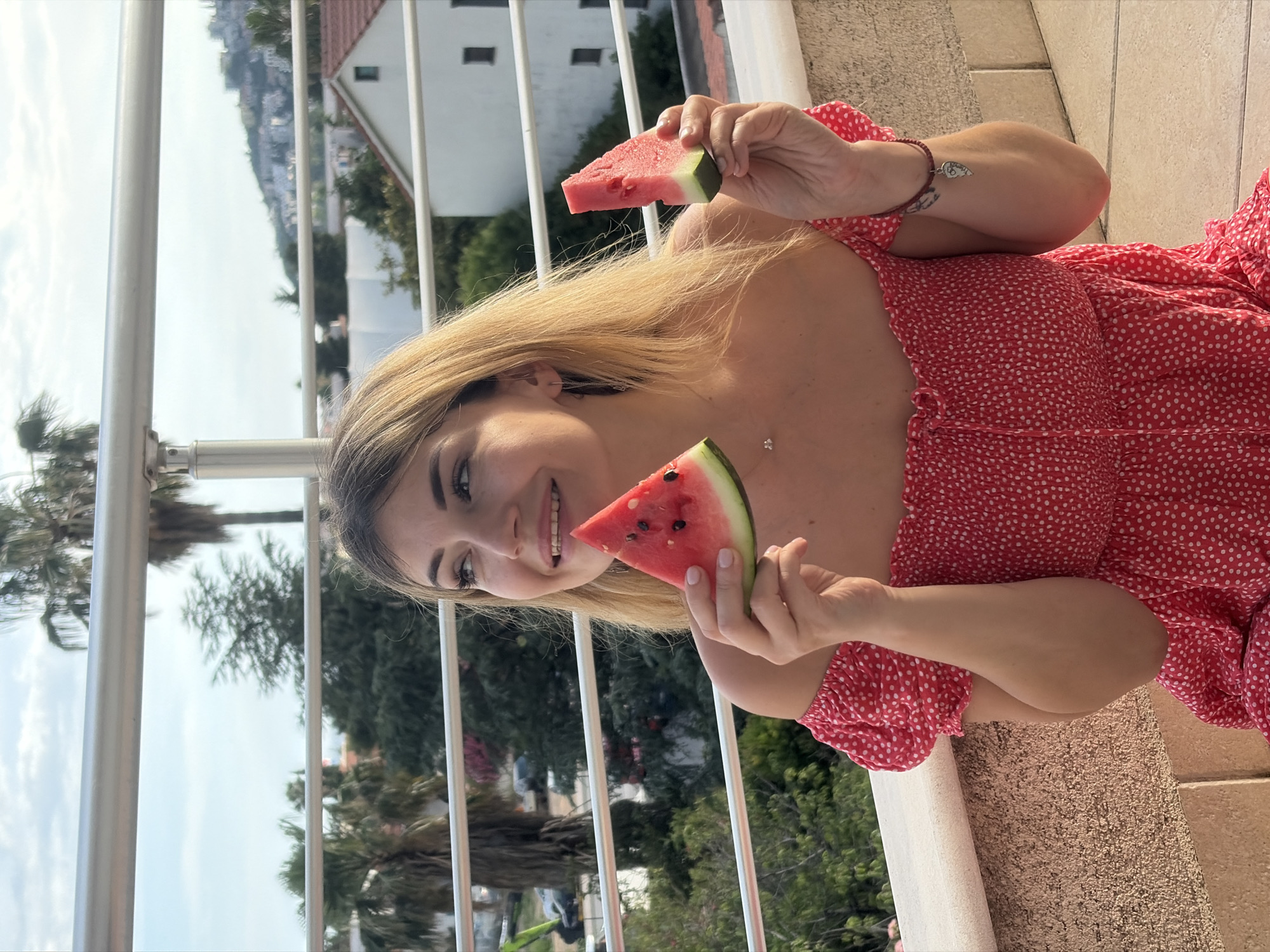 The image displays a female individual, approximately 25-35 years old, positioned on a balcony or terrace. She has light blonde, shoulder-length hair and fair skin, with a smiling expression directed towards her right. She is wearing a red off-the-shoulder dress adorned with white polka dots and featuring a smocked bodice. In her left hand, she holds a triangular slice of watermelon exhibiting dark seeds, and in her right hand, she holds a smaller, more rectangular slice of watermelon. Accessories include a subtle necklace with a small pendant, a stud earring in her left ear, and a red bracelet with a silver heart-shaped charm on her right wrist.
The immediate foreground includes a white metal railing and a textured light-brown wall. Beyond the railing, the midground features a variety of green foliage, including discernible palm trees and other dense trees. Several buildings are visible, most notably a white house with a red-tiled roof to the right and other white structures further in the distance. Partially obscured by trees on the left, an excavator or similar heavy machinery is present. The background shows a light blue sky with scattered white clouds, giving way to a distant hillside densely populated with numerous white buildings, characteristic of a town built on an incline. This urban and natural landscape, featuring Mediterranean architecture and flora, is consistent with coastal regions, specifically within the city of Budva, Montenegro.