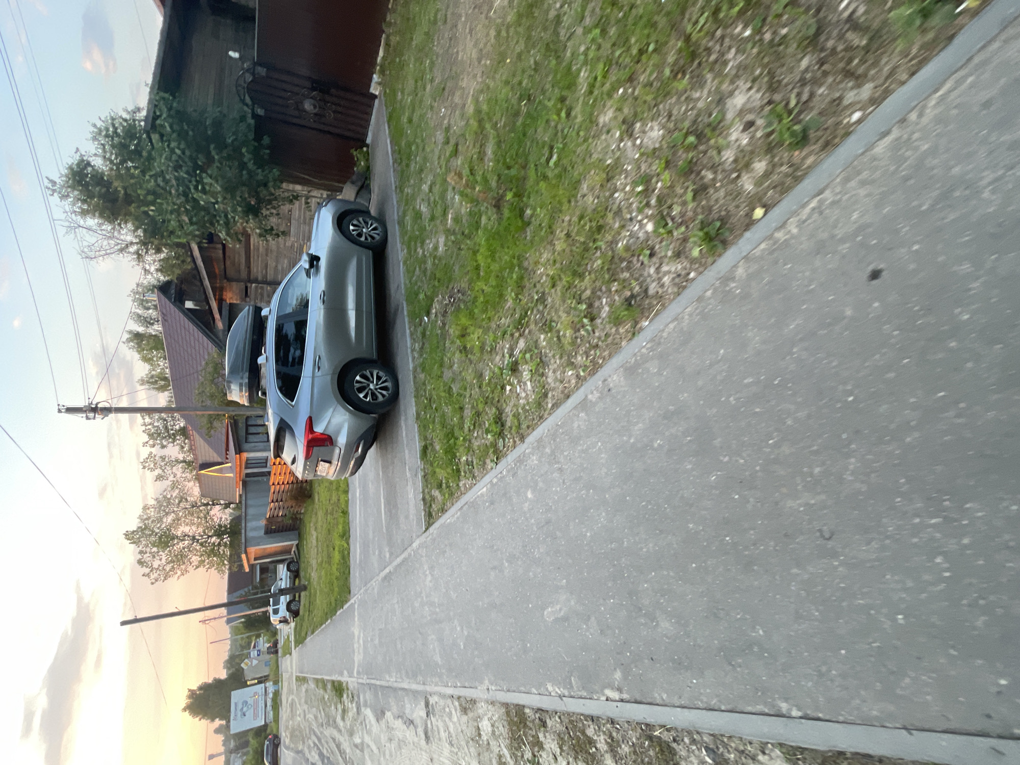 The image depicts a street scene in the village of Krasnye Baki, Russia, during daylight hours. A silver SUV with a roof cargo box is parked on a paved driveway beside a grassy area. To the left of the SUV, a narrow concrete sidewalk runs along a dirt shoulder. In the background, residential buildings with wooden fences and mature trees are visible. The sky overhead is partly cloudy with a soft, diffused light, suggesting it might be late afternoon or early evening. The overall environment appears to be a quiet, residential area.