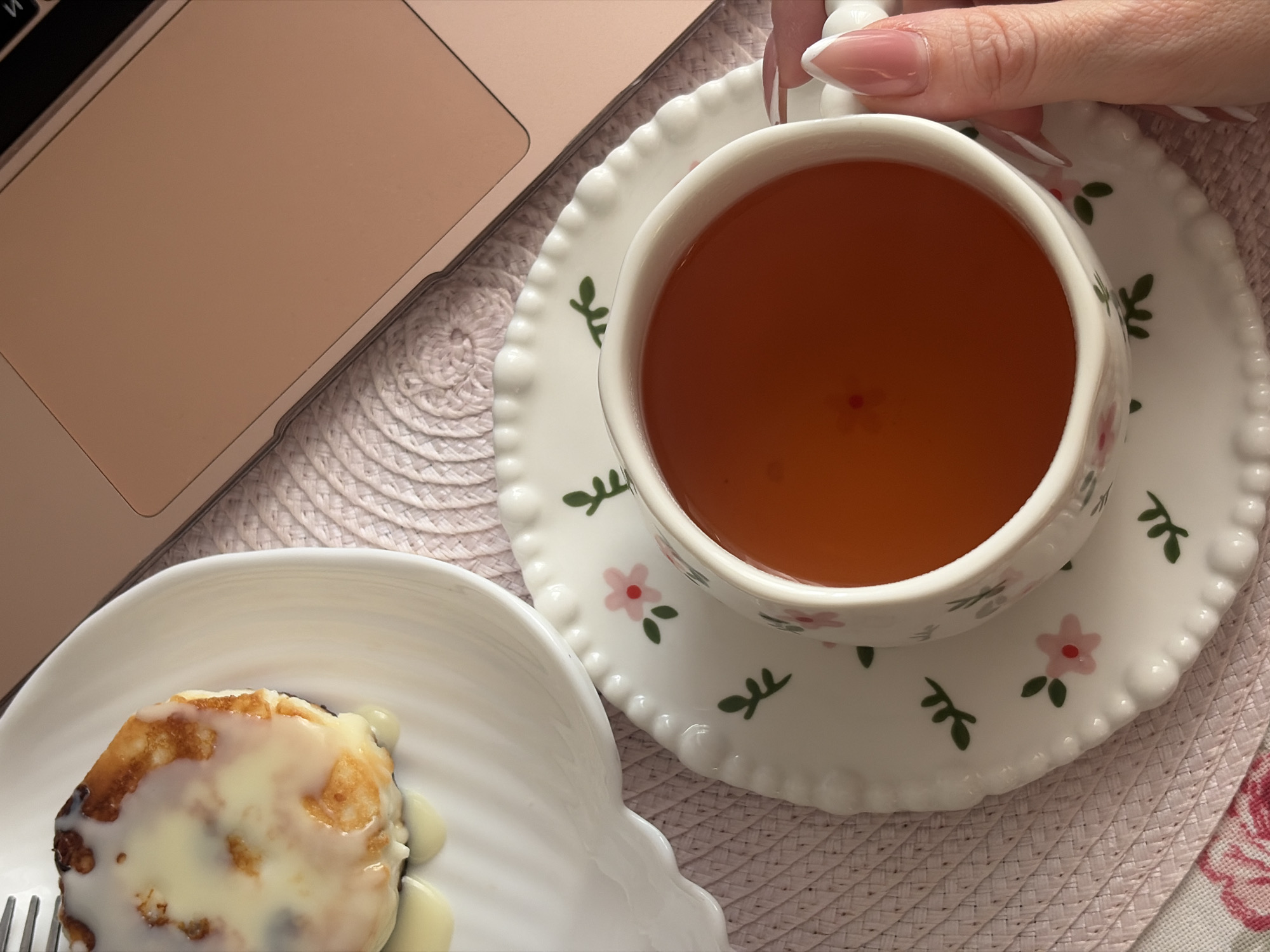 The scene is set indoors in Saint Petersburg, Russia. A close-up, overhead view depicts a tabletop arrangement of breakfast or a snack alongside a laptop. In the foreground, a female hand with a French-tip manicure holds the handle of a white porcelain teacup, which is adorned with a pattern of pink flowers and green leaves. The teacup is filled with an amber-colored liquid, presumed to be tea, and rests on a matching saucer. To the left, a white, shell-shaped plate holds a single, round, golden-brown fried pastry, heavily coated in a thick, white, viscous glaze that has dripped onto the plate. Beneath the teacup and plate is a light pink, circular, tightly woven placemat. In the upper right quadrant, a portion of an open, rose-gold colored laptop is visible, displaying a section of its black keyboard. A small segment of a white fabric with a pink and red floral pattern is visible in the lower left corner. The environment appears to be a casual setting, possibly a home or cafe.