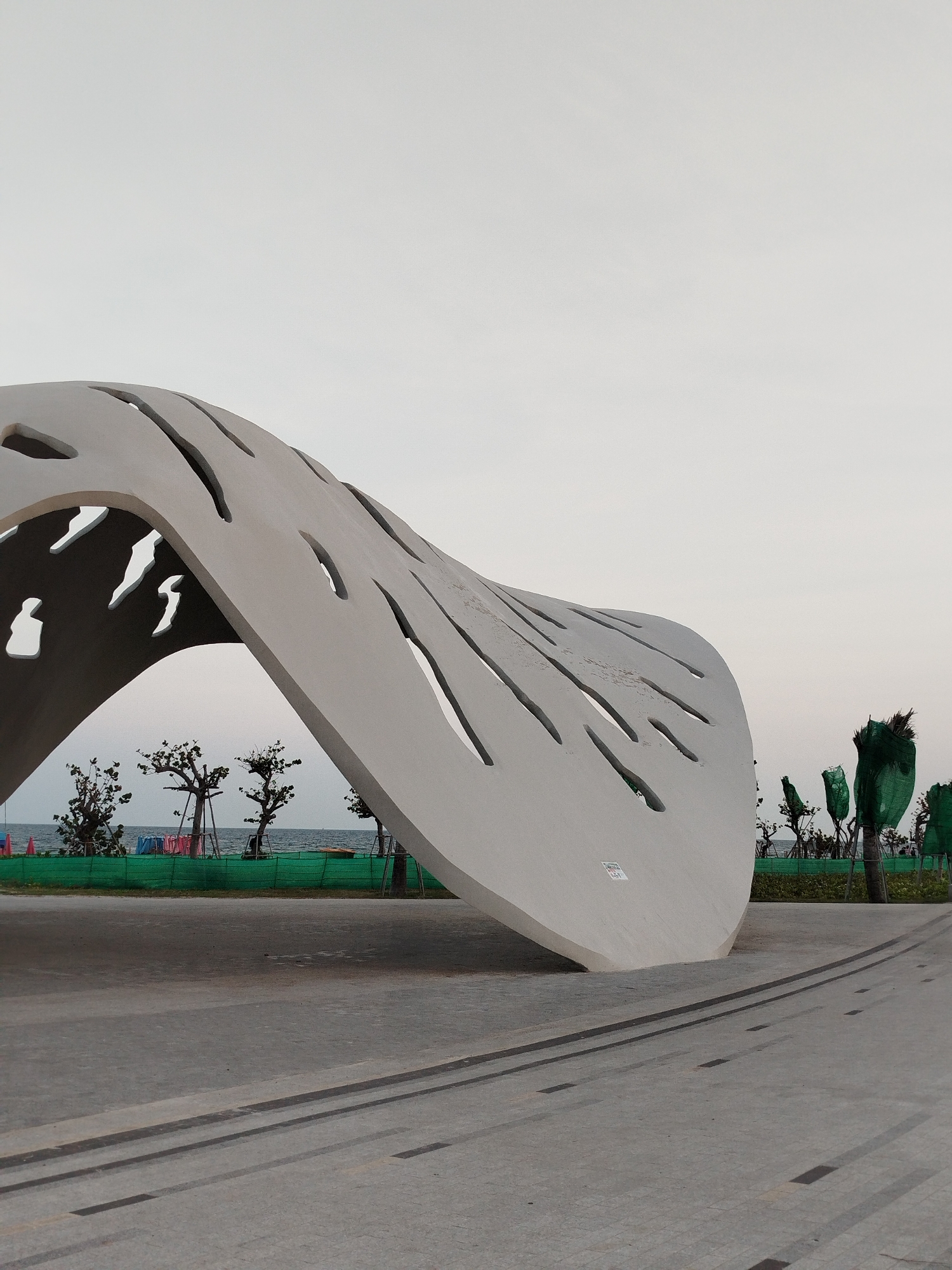 An eye-level, full outdoor shot depicts a large, gray, undulating sculpture in a park-like setting, possibly in Ho Chi Minh City, Vietnam, though the specific location cannot be confirmed.
The sculpture is the focal point, constructed with smooth, gray material, possibly concrete. It has elongated, organic-shaped cutouts across its surface. The structure appears to be both modern and abstract.
Behind the sculpture, the scene opens to reveal a distant shoreline with a beach. Between the sculpture and the shore, a low, green fence borders the area. Several small trees line the fence, some wrapped in green mesh.
The foreground is a paved surface with dark, recessed lines that create a patterned design. The light is diffused, creating a soft, overcast ambiance. The sky is a pale, uniform color, suggesting an overcast day or early evening. There are no people visible in the image. The overall impression is one of calm and tranquility in a well-maintained public space.