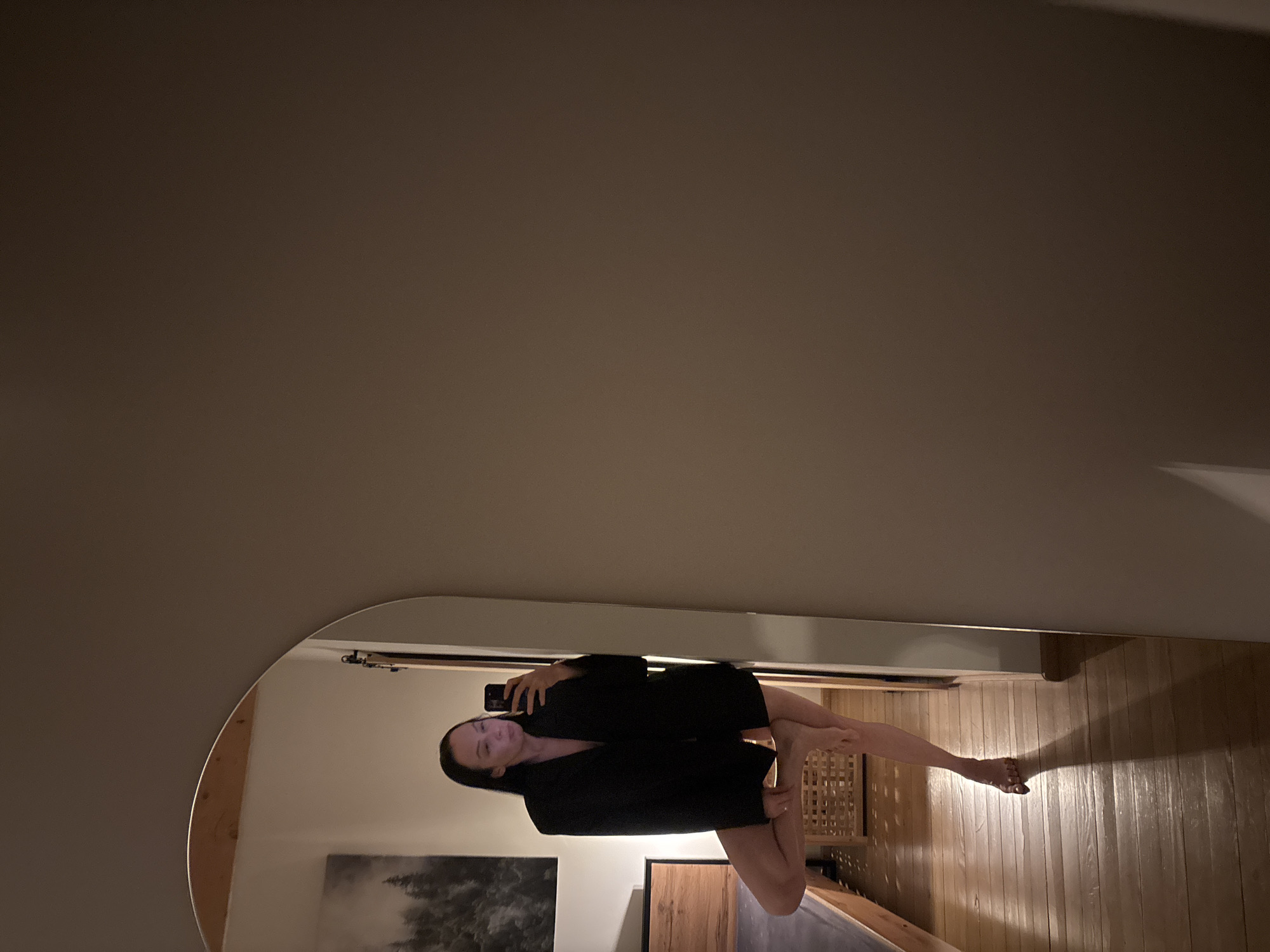 The image depicts an indoor scene in Chaikovsky, Russia, under dim lighting conditions. A large, arched mirror on the left side of the frame reflects a female individual. She possesses long, dark hair and is clad in an oversized black blazer, with her lower body uncovered. The individual stands in a single-leg pose, with her right foot resting on her inner left thigh; her left foot is shod in a light-colored, open-toed sandal. She holds a dark smartphone in her right hand, positioned to capture her reflection, with a faint purple light observed on the device's screen. The mirror's reflection reveals a wooden bed frame with dark bedding to the subject's left, a framed landscape print depicting dark trees on the wall above the bed, and a light-colored woven basket positioned on the wooden plank floor. A distinct shadow from the subject is cast onto the floor. The right side of the image displays a plain, light-brown wall.