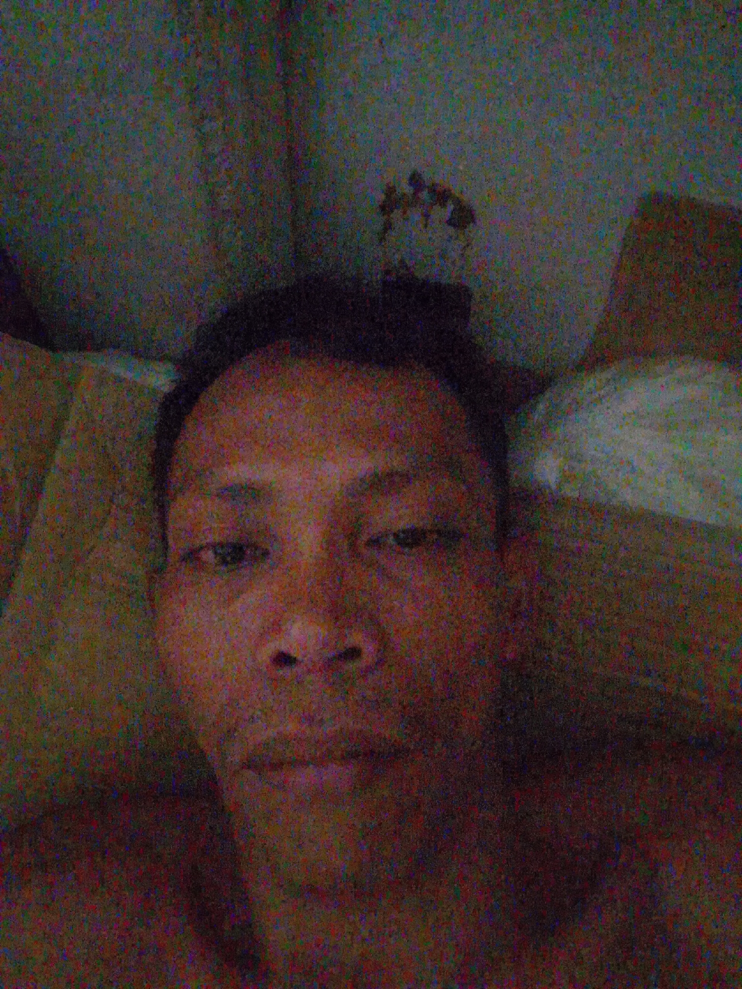 The image shows a close-up selfie of a man with dark hair. He appears to be lying down, possibly in a bed, with a pillow or bedding visible behind him. The background is unclear due to image noise, but a wall is discernible. The scene is presumed to be located in Bukit Ketok, Indonesia, based on the prompt.