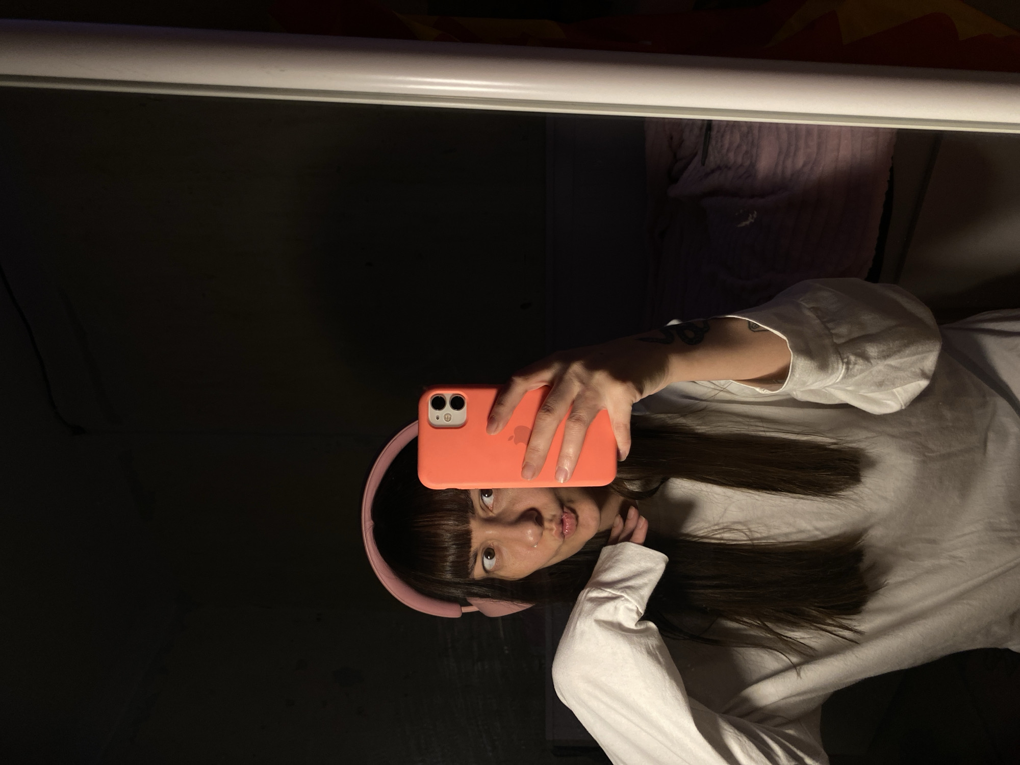 A young adult is reflected in a mirror, positioned indoors. The person is holding an orange iPhone in their right hand, presumably taking the picture. They have dark hair with bangs, a lip piercing, and are wearing pink headphones and a white long-sleeved shirt. The background includes a dark wall, a white horizontal frame (presumably part of the mirror or a nearby structure), and a glimpse of bedding. The location cannot be determined as Votkinsk, Russia, with only the provided image.