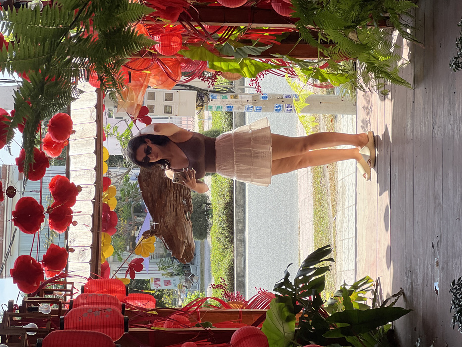 A woman wearing a brown tank top and white skirt poses in front of a decorative display. Red paper lanterns and various floral arrangements frame the foreground. She holds a phone, seemingly taking a photo of herself. Sunlight streams in, illuminating the wooden floor and a view of the street and greenery outside. The perspective of the photo is rotated and slightly angled.