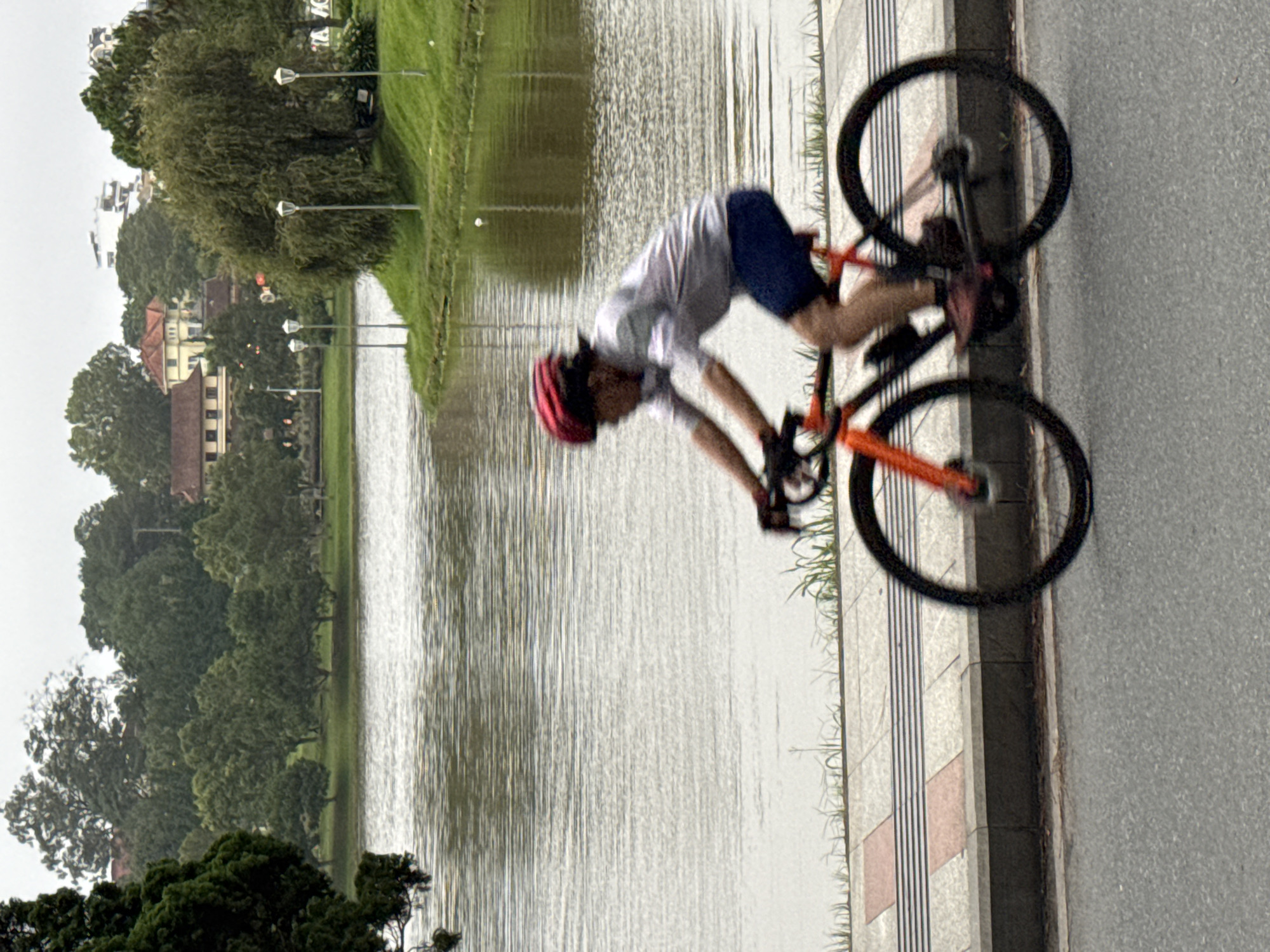 The image shows a person riding a bicycle on a paved path beside a body of water. The rider, wearing a red helmet, light-colored short-sleeved shirt, and dark shorts, is in motion, indicated by the blur of their body and the bicycle's wheels. The bicycle is orange and black. The path is bordered by a concrete curb and grass. In the background, across the water, is a tree-lined landscape with buildings and lamp posts visible, suggesting an urban park or scenic area. The water is calm, reflecting the overcast sky. This scene is consistent with the environment around Xuân Hương Lake in Đà Lạt, Vietnam, known for its cycling paths and picturesque surroundings.