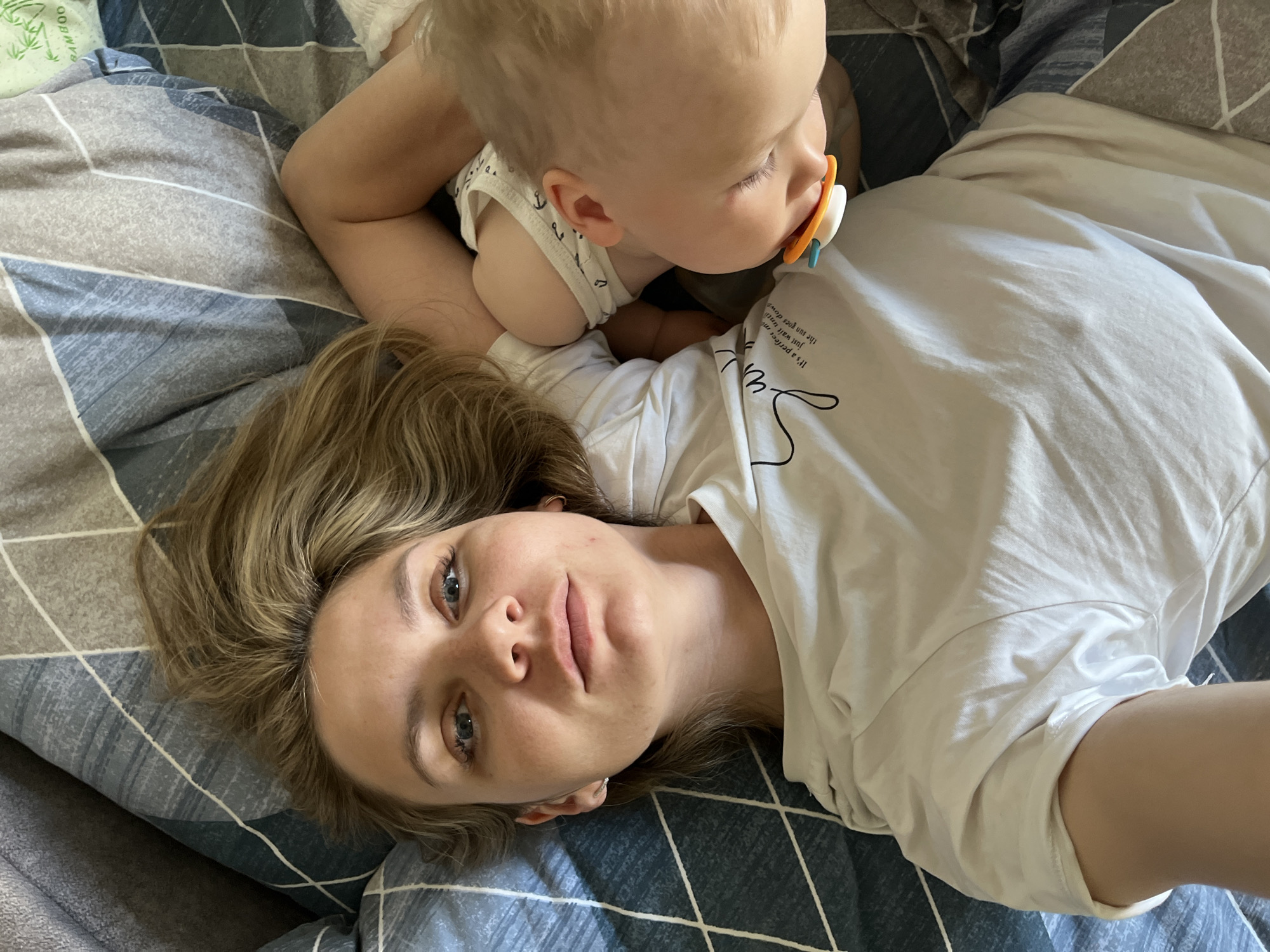 A woman and a baby are lying on a bed with a blue and grey patterned comforter. The woman has blonde hair and blue eyes, and she is wearing a white t-shirt. The baby is wearing a white tank top with small anchor designs and has a pacifier in their mouth. The woman is looking at the camera, and the baby is looking to the right. The location is likely indoors, specifically a bedroom, though the image offers no evidence to suggest this is Chaikovsky, Russia.