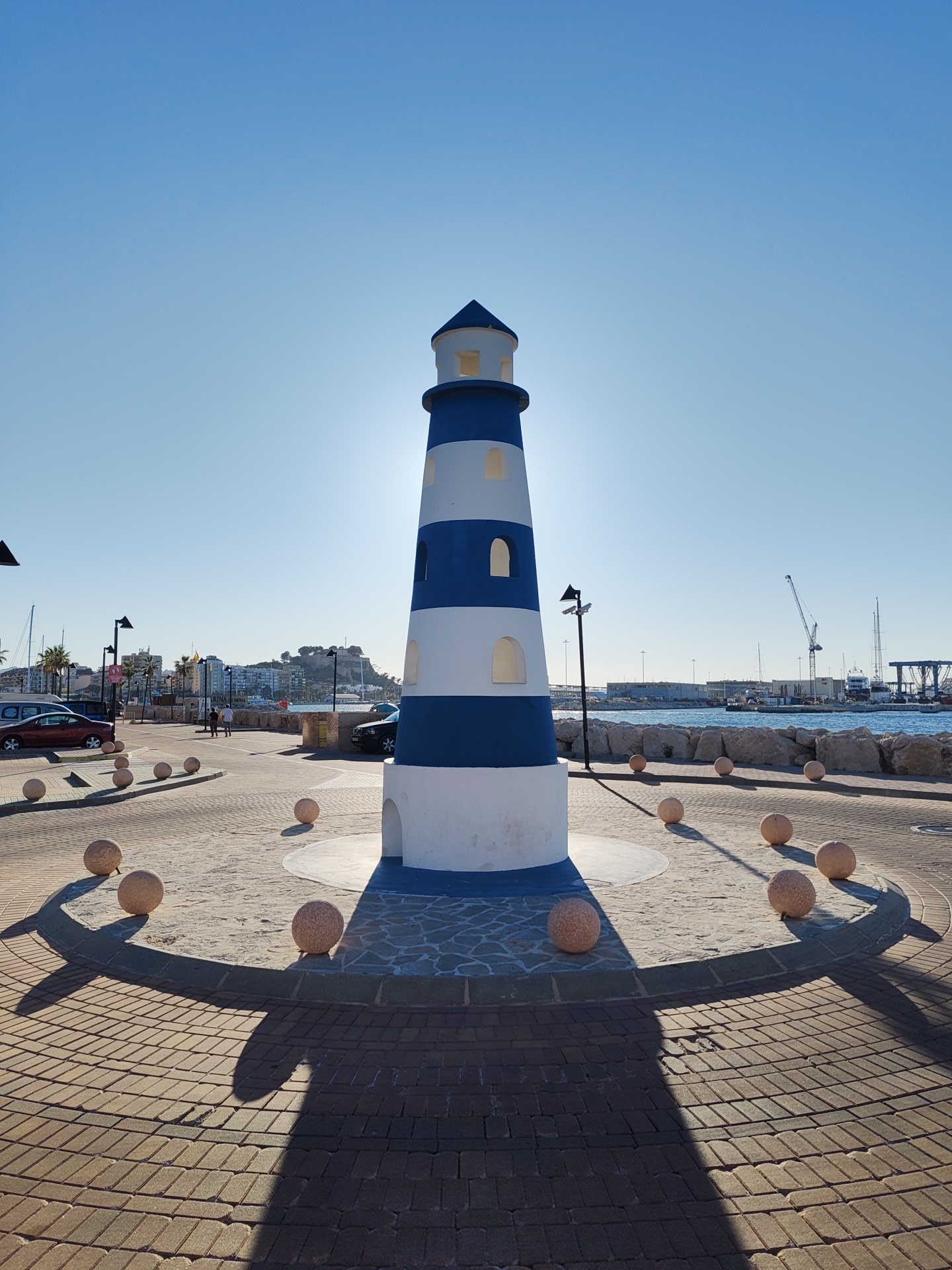 The image captures a clear, sunny day in Dénia, Spain. In the foreground stands a decorative lighthouse, painted with alternating blue and white horizontal stripes. The structure is centrally positioned on a circular platform, with round, stone-like balls arranged around the perimeter. The ground surrounding the lighthouse is paved with brick, and the long shadow of the lighthouse stretches across the pavement. In the background, a coastal scene includes the sea, a harbor with cranes and boats, and a cityscape with buildings and hills. A few cars and lampposts are visible near the lighthouse. The overall environment appears to be a public space, possibly a promenade or roundabout.
