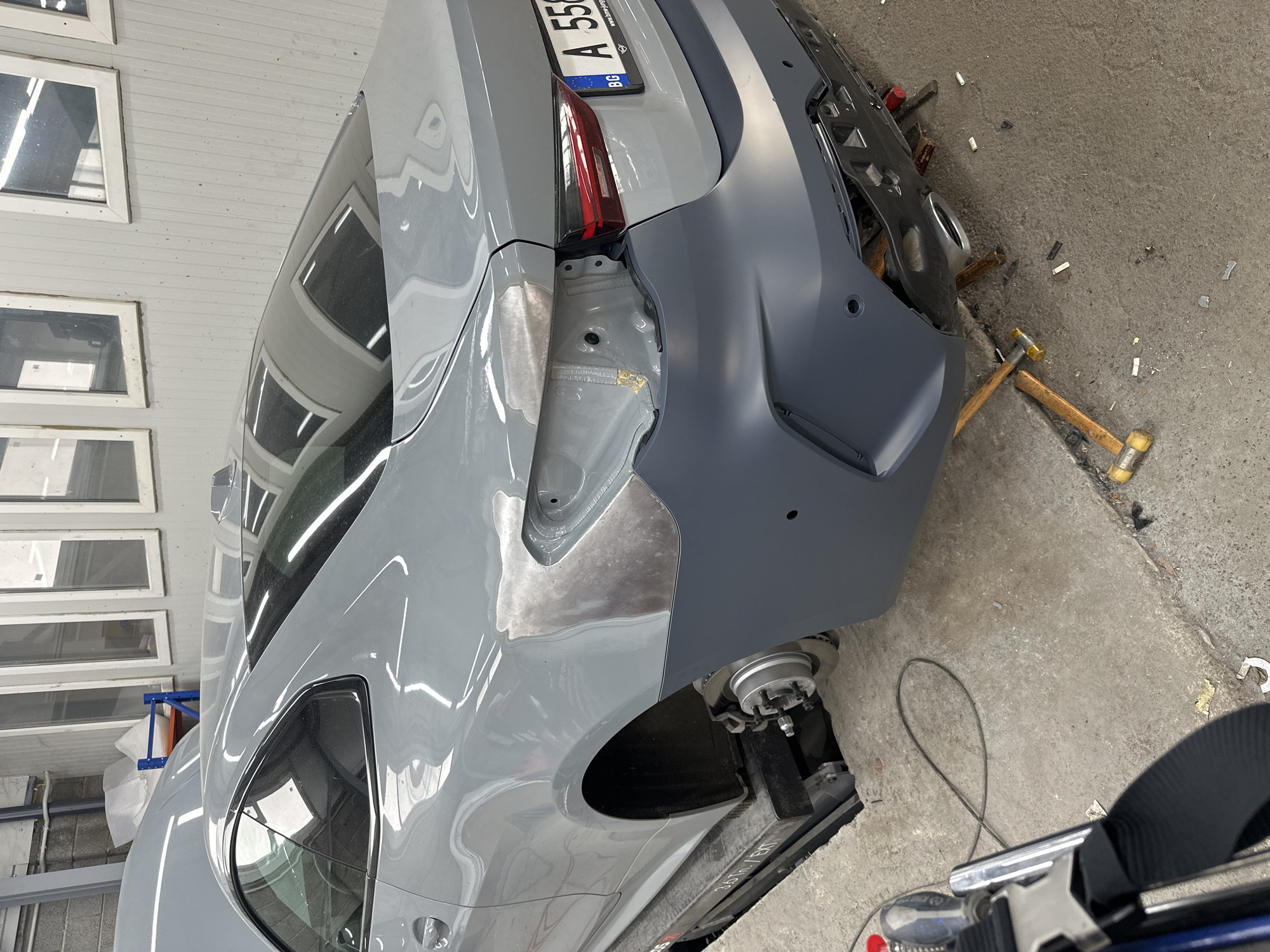 An indoor scene captures the rear right quarter of a gray car undergoing body repair in an auto body shop or garage. The vehicle, a modern sedan or coupe, is elevated on a hoist, with its rear wheel removed and suspension components visible.
The car's rear right quarter panel shows areas of exposed metal or body filler, indicating active repair work such as dent removal and panel shaping. A dark gray, primed bumper, likely a new or repaired part, is partially fitted to the rear, covering some of the internal structure. The car’s original gray paint is visible on the remaining body panels, which reflect the surrounding environment, including the rectangular wall features. A red taillight is visible on the left side of the rear, and a Bulgarian license plate (BG A 550) is mounted on the trunk lid.
On the concrete floor beneath the car, several tools are scattered, including two hammers with wooden handles and yellow/white rubber or plastic heads. Small white debris pieces are also present. A black electrical cable forms a loop on the ground. In the background, light gray walls feature multiple rectangular openings, some with glass, reflecting light. A blue and silver structure is partially visible on the left. Text on the car lift arm reads "3.6T / EXT". No individuals are present in the image. The scene depicts a vehicle in an advanced stage of body repair, being prepared for subsequent finishing or painting.