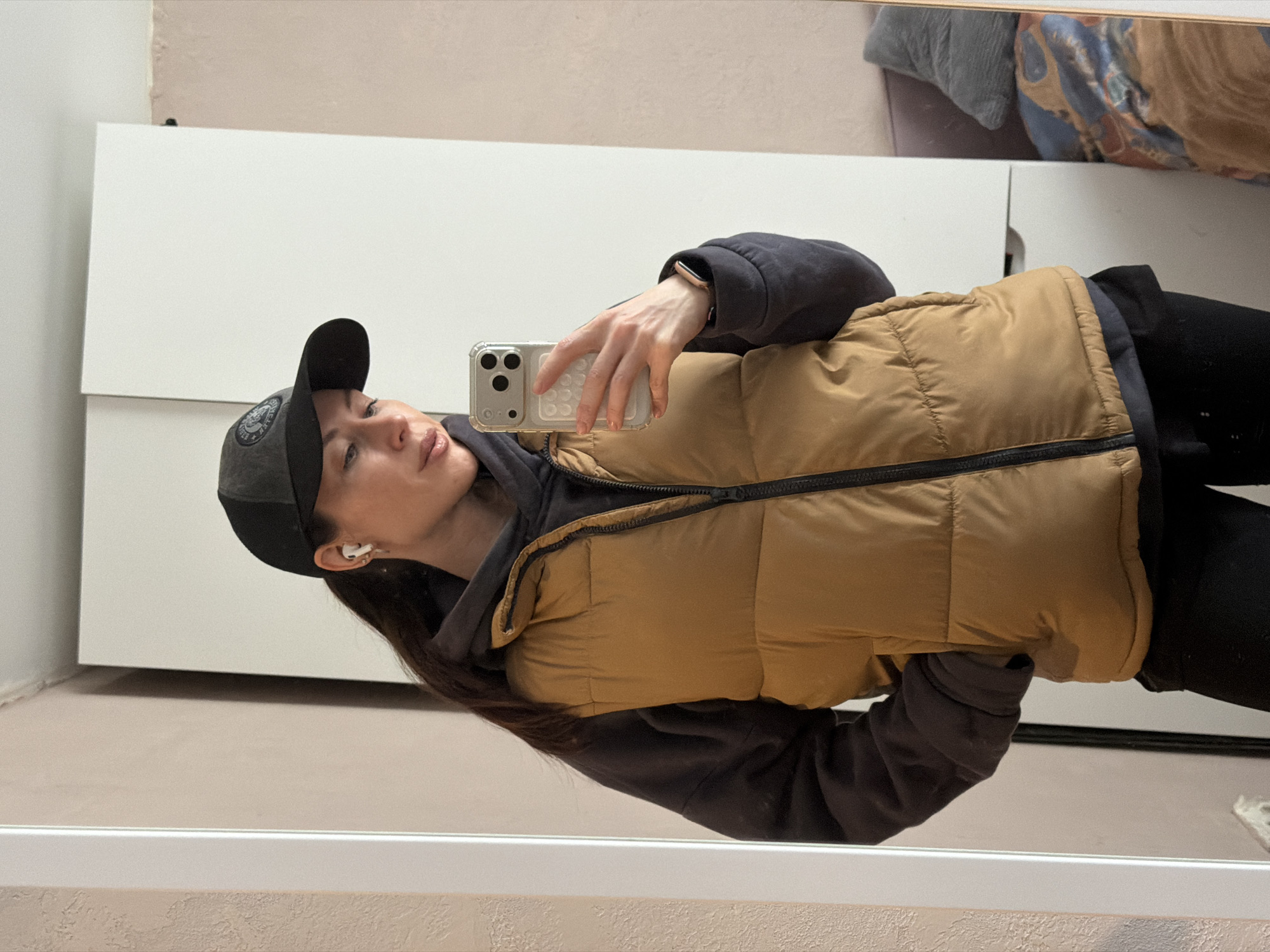 This vertical mirror selfie, taken in Perm, Russia, features a young woman dressed in casual attire. She is positioned horizontally within the frame, looking towards her smartphone which she holds up to take the picture. She wears a dark grey baseball cap with an indistinct circular patch or logo on the front. Over a dark hooded sweatshirt, she sports a tan or camel-colored puffer vest, paired with dark trousers. White wireless earbuds are visible in her right ear, and a light-colored smartwatch adorns her left wrist. The smartphone she uses, likely an iPhone due to its camera module, is light-colored and protected by a clear, textured case.
The background, reflected in the mirror, indicates an indoor setting, likely a bedroom or dressing area. A large white two-door wardrobe or cabinet occupies the left side of the reflection. Above it, a light beige wall shows some minor wear, including what appears to be peeling paint. In the upper right portion of the reflection, elements of a bed, including a grey pillow and a blue and orange patterned blanket, are visible. The floor beneath the wardrobe is a uniform light beige. The lighting suggests it is daytime. The woman's expression is neutral, focused on her device. No discernible text is clearly legible within the image.