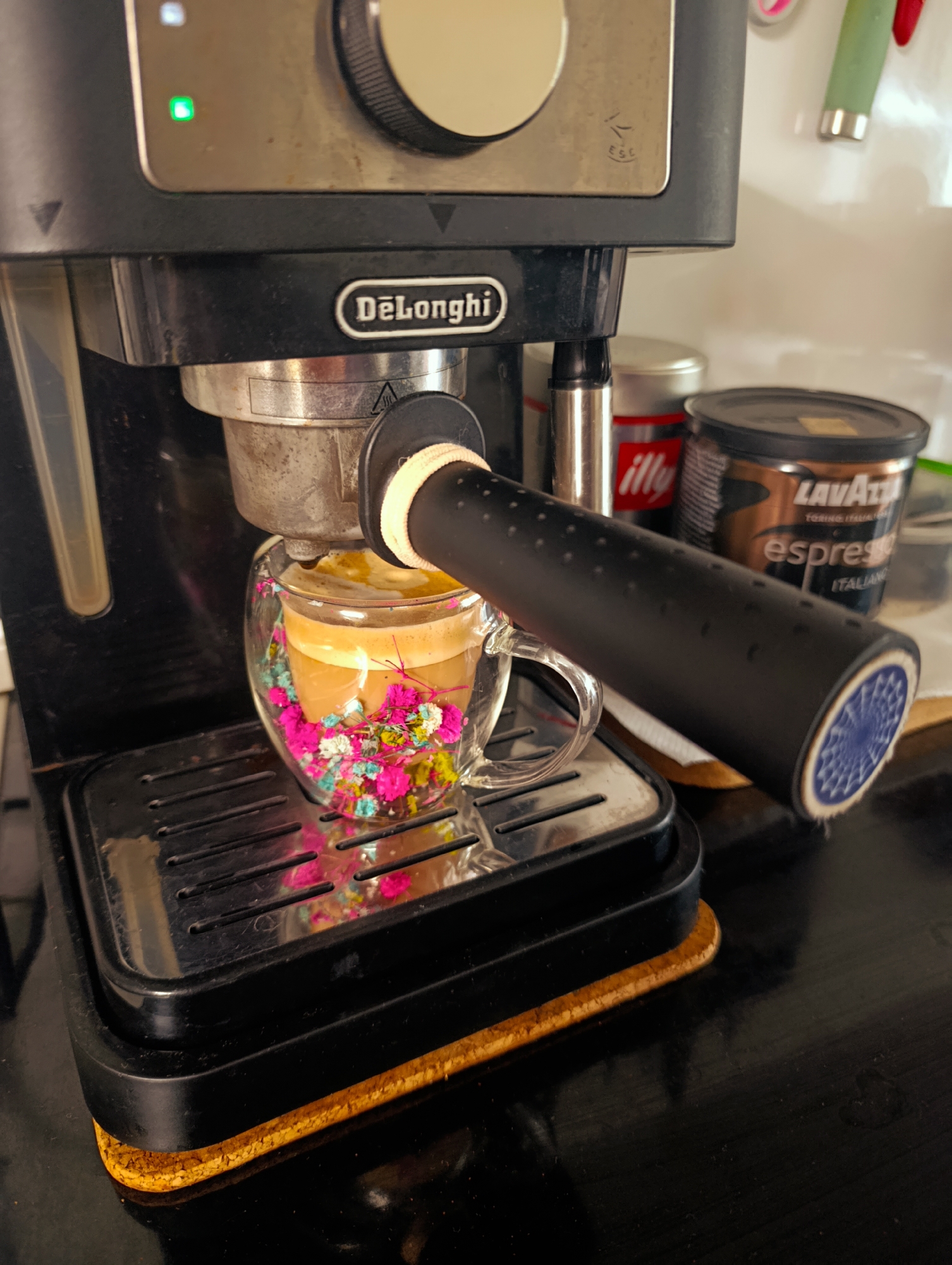 A close-up shot depicts a black DeLonghi espresso machine in operation. The machine is positioned on a black surface with a cork coaster underneath. A clear double-walled glass cup with decorative flowers at the bottom is placed beneath the portafilter, catching the freshly brewed espresso. To the right of the espresso machine, partially visible, are a tin of Illy coffee and a brown Lavazza espresso tin. The background is out of focus, showing a white wall with some partially visible objects. The location is not identifiable as Kardzhali, Bulgaria, from the provided image.