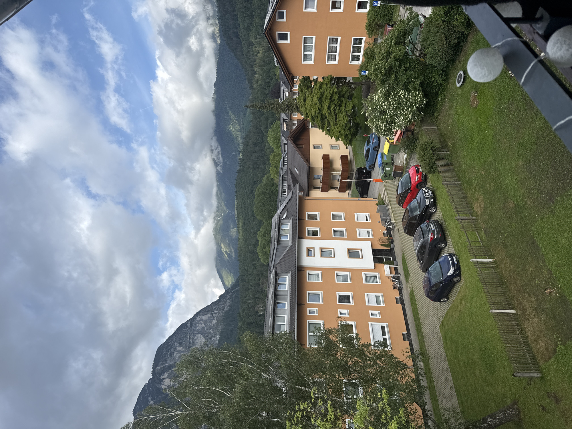 This image depicts a residential area in the town of Garmisch-Partenkirchen, Germany, set against a mountainous backdrop. The scene is observed from a slightly elevated vantage point, likely a balcony, as indicated by a dark railing with decorative spherical lights in the bottom right corner.
The sky is predominantly overcast with varying shades of grey clouds, though patches of blue sky and white cumulus clouds are visible, particularly towards the right side of the frame. In the background, large, dark mountains dominate the horizon. Their lower slopes are densely covered with dark green coniferous trees, while higher elevations reveal rocky outcrops and are partially obscured by low-hanging clouds or mist.
In the mid-ground, a cluster of multi-story residential buildings is visible. The prominent buildings in the left and right foreground are uniformly painted in an orange-terracotta hue with white window frames and dark grey pitched roofs. The building on the left features an attic level with multiple dormer windows. Another building further back, positioned centrally, exhibits a light brown facade with dark roofs and balconies. The architectural style appears to be modern alpine.
Between and around these buildings, several vehicles are parked on paved parking lots made of interlocking grey pavers. In the immediate foreground, a dark blue hatchback is parked. Behind it, a dark grey SUV is visible. Further left, a red compact SUV and a black SUV (partially obscured) are parked. A blue sedan is parked on a paved area further back on the left, next to green and yellow waste bins. A black van is partially visible behind the blue sedan. Blue parking signs with a white 'P' are present in various locations.
The landscape includes significant vegetation. A large deciduous tree with white bark and green leaves stands prominently on the left, partially obscuring the mountains and one of the buildings. A well-maintained grassy lawn with a low wooden picket fence separates the immediate foreground from the parking areas and buildings. A large tree with white blossoms is visible near the center-right, surrounded by shrubs. A small, dark green conifer stands centrally between some of the buildings. On the grassy area in the mid-right, a small, circular trampoline-like object is present.
No people or discernible human actions are depicted within the frame. The overall impression is one of a quiet, residential setting within a scenic mountainous environment.