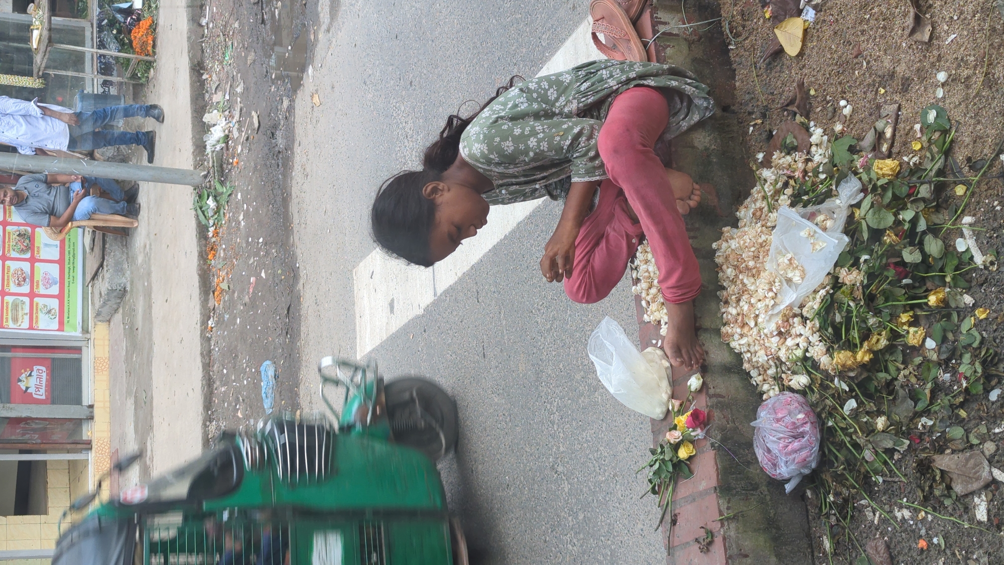A young girl with dark hair, wearing a green floral print dress and pink leggings, sits cross-legged on a raised concrete curb beside a street in Dhaka, Bangladesh. She is looking down at her hands, which are holding small white flowers, and appears to be arranging them. In front of her is a large pile of discarded flowers, some fresh and some wilted, along with two plastic bags, one of which is filled with white flowers. To her left, several fresh roses in red, yellow, and pink hues are laid out. A pair of brown sandals rests on the curb behind her. In the background, a green auto-rickshaw is in motion on the asphalt road, and a few people are visible on a sidewalk near buildings, one of whom is seated on a chair. The ground around the flowers is a mix of dirt and debris.