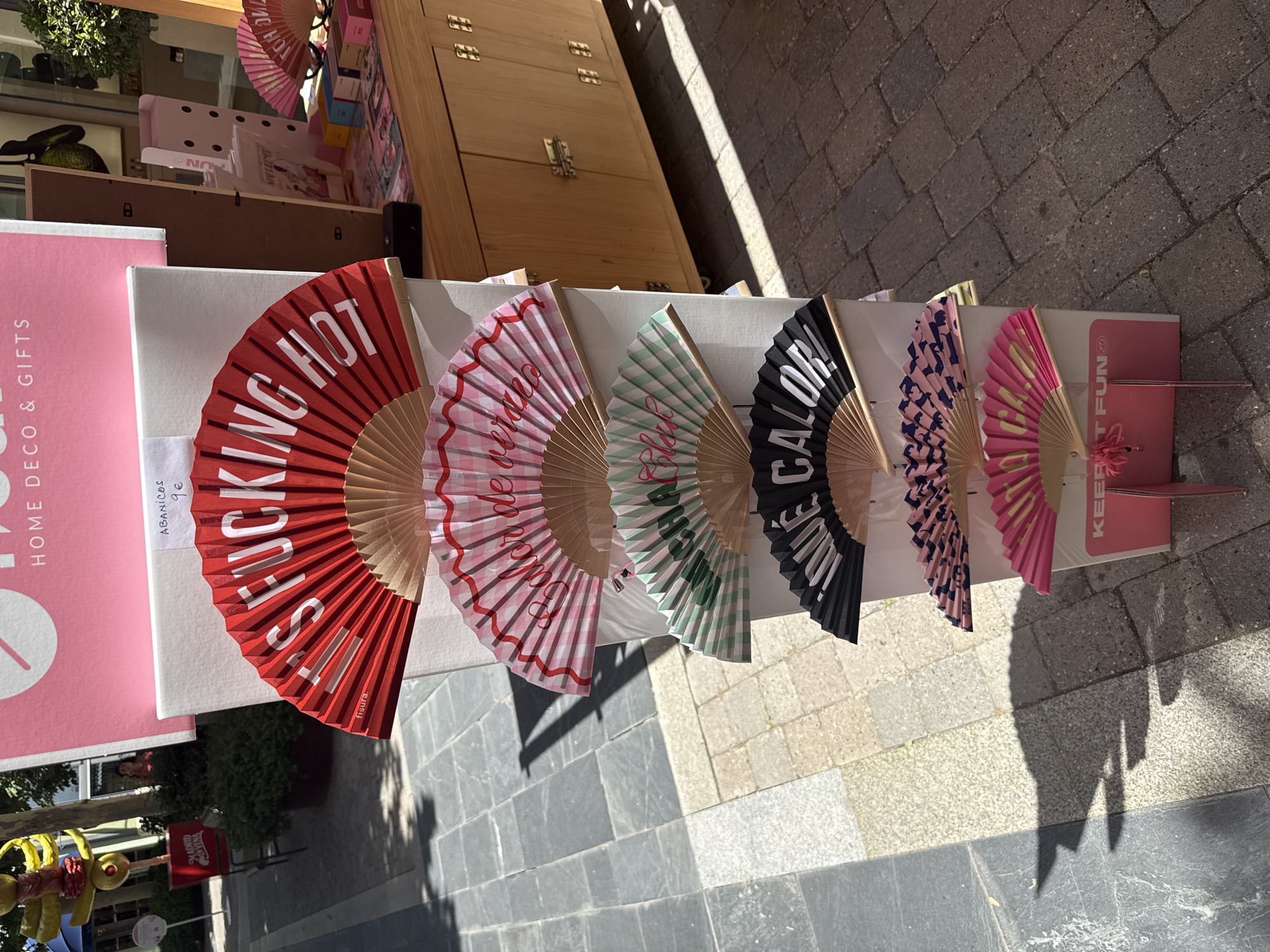 In Las Rozas de Madrid, Spain, an outdoor market stall displays several vibrantly colored folding fans. The fans are arranged vertically, fanned open to reveal various text and patterns. The top fan is red with white text that reads "FUCKIN' HOT". Below it, a pink and white checkered fan displays the text "Calor de veran" (Summer heat). A green and white checkered fan follows, with the text "Clak!". The next fan is black with white text that says "MUCHO CALOR!" (Very hot!). The bottom two fans feature patterned designs, one with a navy blue and pink geometric pattern, and the last one is bright pink with yellow text. A sign at the bottom reads "KEEP IT FUN". The stall is set up in front of a wooden cabinet, and the ground is paved with grey and brown square tiles, casting strong shadows from the sunlight. In the background, a large pink banner with white text "HOME DECO & GIFTS" is visible.