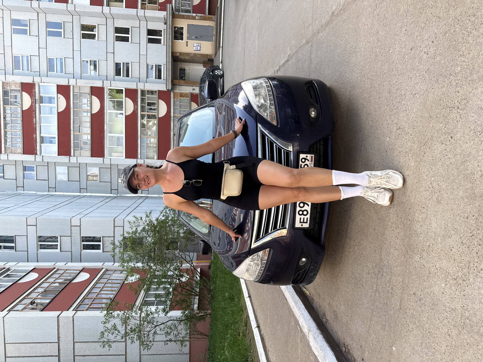 The image captures a young woman in Chaikovsky, Russia. She is posing with a dark blue car, leaning against its hood. The car's license plate is visible, reading "E89." The woman is wearing a black tank top, black shorts, white socks, and white sneakers. She has a small white purse and a black baseball cap. The backdrop features a multi-story building with a gray facade and red accents. Another car is partially visible in the background. The scene is set on a paved area, possibly a parking lot or street.
