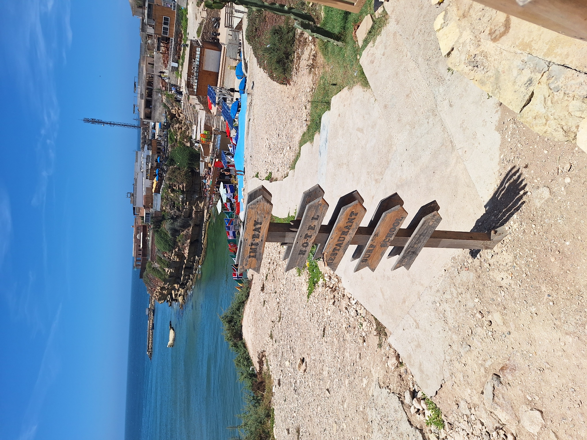 The image shows a wooden signpost in the foreground with multiple directional arrows indicating "IMI-BAY", "HOTEL", "RESTAURANT", and "SURF SCHOOL". The signpost stands on a dusty, rocky path that leads down concrete steps. In the background, there is a coastal village on a cliff overlooking the ocean. Several buildings, including what appear to be hotels and restaurants, are visible. A bright blue swimming pool is situated on a terrace, with people lounging around it. The ocean is calm and blue under a clear sky with a few wispy clouds. The overall scene depicts a tourist destination in Imsouane, Morocco, likely a coastal resort area known for surfing.