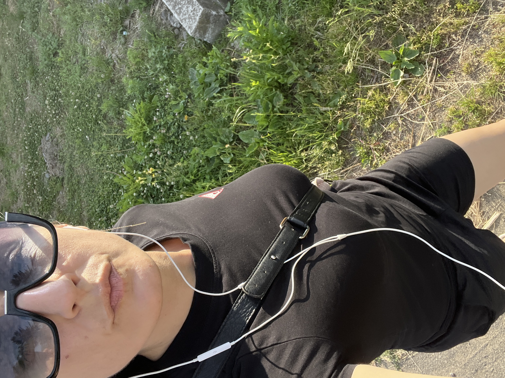 The image depicts a person, likely female, in Tarnivtsi, Ukraine. The subject is shown from the upper torso to mid-thigh, viewed from a slightly elevated, close-up perspective. She wears large, black-framed sunglasses, reflecting tree imagery, and a black t-shirt with a small, partially visible red/orange triangular emblem on the right chest. White wired earbuds are inserted into both ears, with the wire running over her chest and a control module visible. A black, textured strap, possibly from a bag, with a metal buckle and rings, crosses her torso diagonally. A single gold-colored dangle earring is present in her right ear. The lower body shows black shorts or a skirt and a bare leg. Her facial expression is neutral with slightly pursed lips. The outdoor environment consists of a grassy area with various green plants, weeds, small white and yellow wildflowers, and patches of bare earth. A concrete block is visible on the far right, and a paved or asphalt surface is in the lower-left foreground. The scene is brightly illuminated by sunlight, casting distinct shadows.