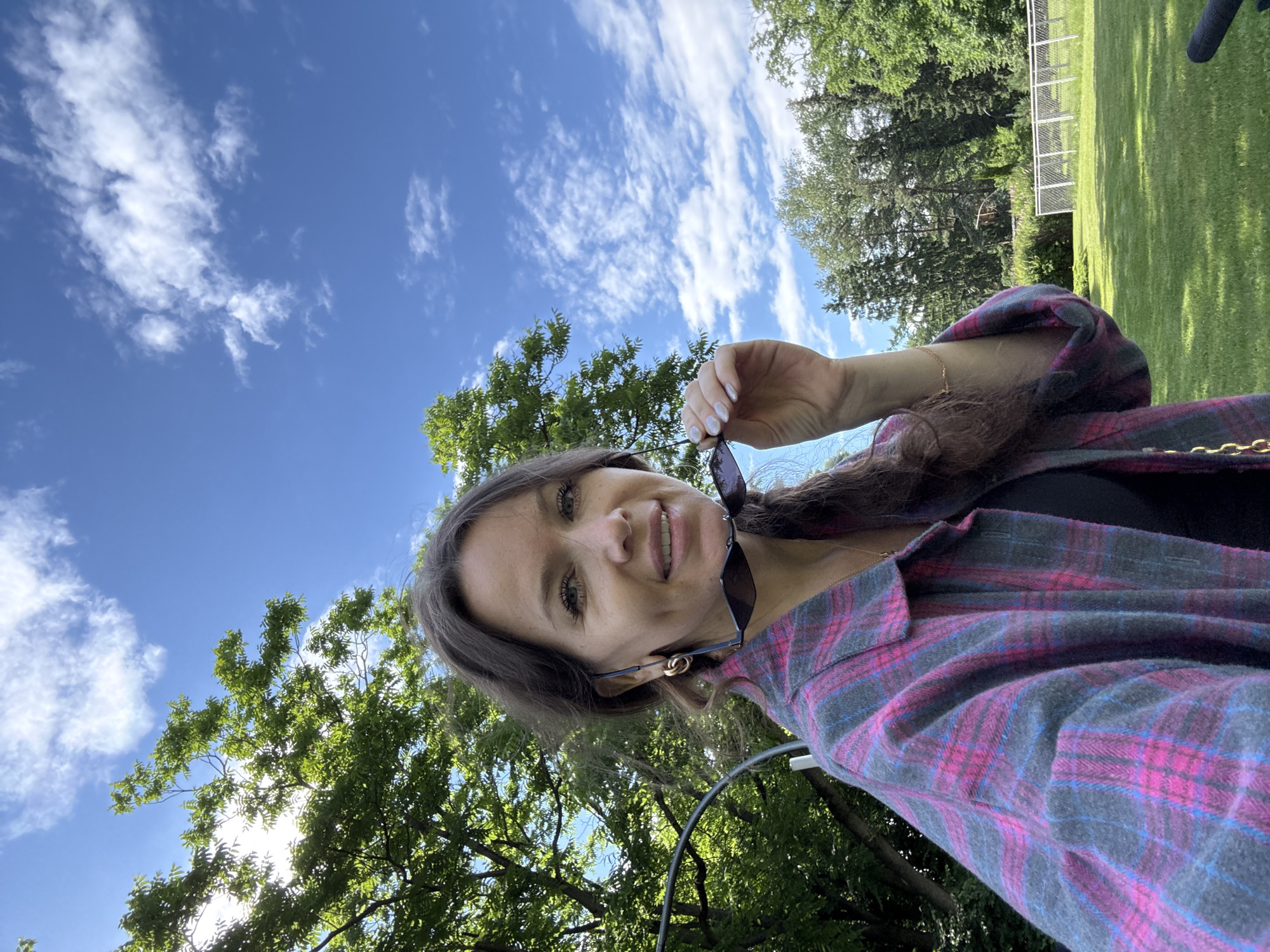 A young woman is taking a selfie outdoors, holding sunglasses in her right hand. She is wearing a pink and grey plaid shirt over a black top and a delicate gold necklace. Her hair is long and dark brown, and she has on earrings with gold loops. The woman has green eyes and a bright smile, conveying a cheerful and relaxed mood. The background is a vibrant green, with trees and a sunny lawn, suggesting a pleasant day. A white fence and what appears to be a sports field are visible in the distance, indicating an outdoor recreational area. The sky above is a brilliant blue with scattered white clouds. The sun is shining, casting shadows on the grass, suggesting it is daytime, likely mid-morning or early afternoon. The overall scene evokes a sense of leisure and enjoyment in a park-like setting. There is no visible text in the image.