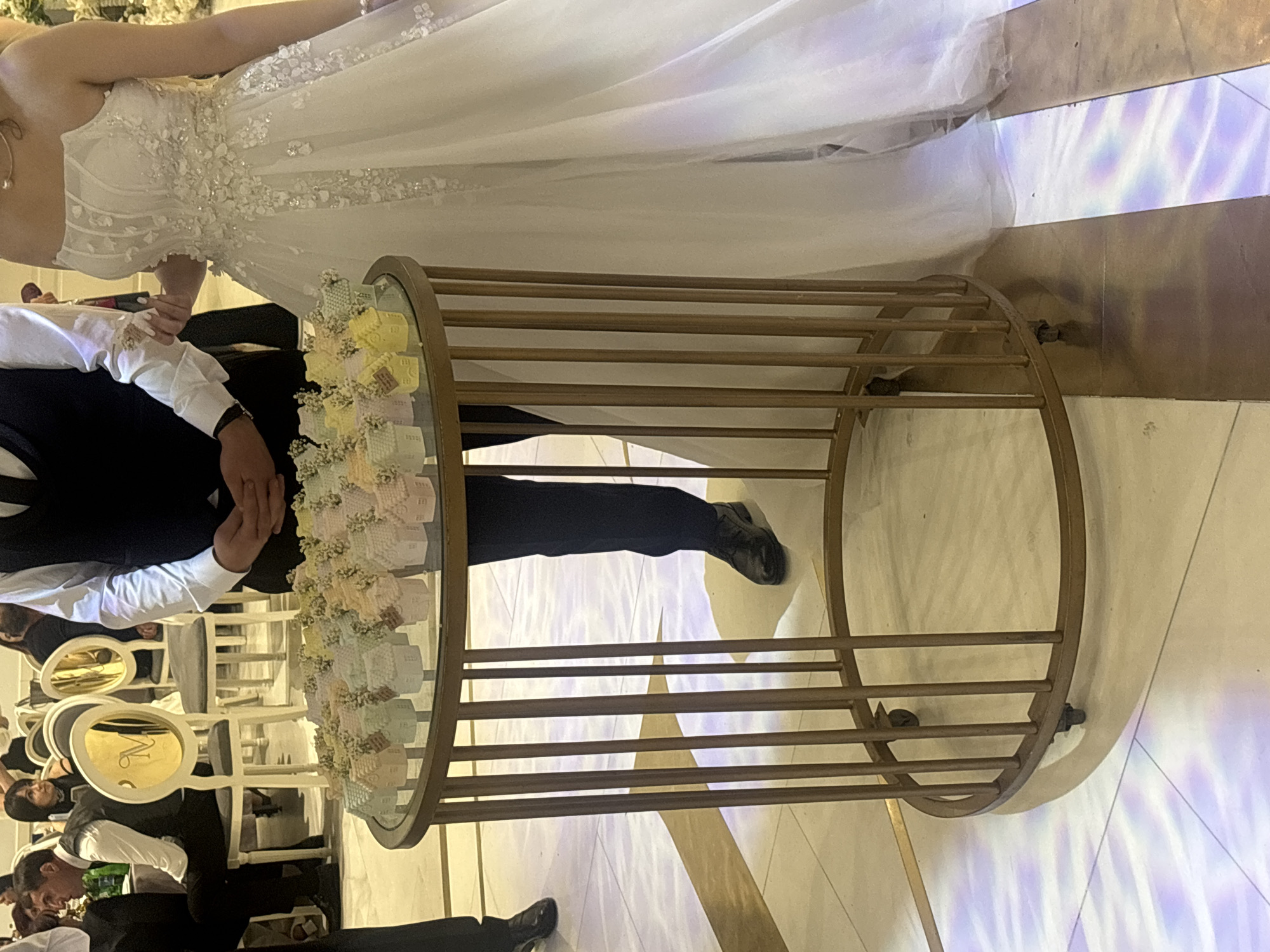 This appears to be a wedding reception scene, possibly indoors, in a location that may be in the city of Masis, Armenia. A bride in a white wedding dress is standing next to a groom wearing a dark suit and white shirt with a bow tie. They are both standing near a round table with a mirror top. The table is surrounded by a metal frame with decorative detailing and is holding small, decorated boxes or favors. The lighting is bright, with a pattern of what appears to be colored light projections, possibly from stage lighting, on the floor. Around the table, other people can be seen seated on chairs.