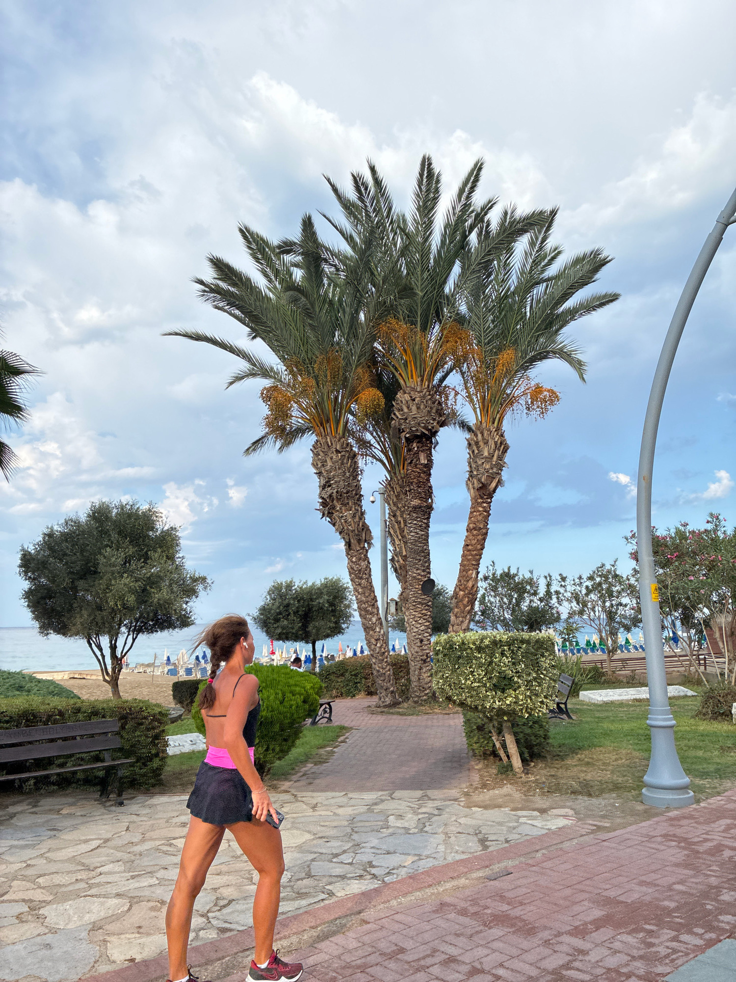 A woman with braided brown hair is walking along a paved pathway. She is wearing a black athletic outfit with a pink waistband and black sneakers. Her right arm is bent, and she is holding a dark smartphone in her hand. She is looking to her right, away from the camera. Behind her, several palm trees with visible fruit clusters are prominent. To the left of the pathway, there are benches and manicured bushes. In the background, beyond the greenery, there is a beach with lounge chairs and umbrellas, and the sea. The sky is overcast with light and dark clouds. The pathway is a mixture of stone pavers and red brick pavers. A large grey lamppost is visible on the right side of the frame. The scene appears to be a promenade or park area in Alanya, Turkey, possibly near the beach.