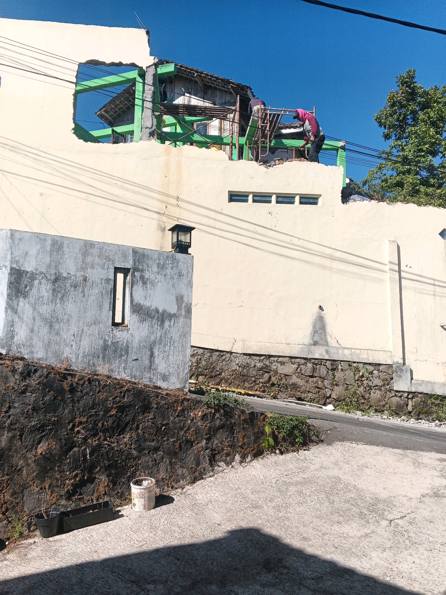 In the city of Keji, Indonesia, a building undergoing demolition or construction is visible against a clear blue sky. Two individuals are working on the upper level, one wearing a pink head covering and the other a dark shirt. They are positioned on scaffolding or a makeshift platform, actively engaged in dismantling or rebuilding a section of the structure. Green metal beams are prominently featured as structural support for the damaged or incomplete part of the building. The building itself has a cream-colored exterior wall with a large, irregular opening revealing the internal construction. A smaller, gray concrete structure with a narrow, vertical window stands in the foreground, partially obscuring the view of the larger building. Electrical wires crisscross the scene overhead, casting shadows. The ground level features a stone retaining wall, some sparse vegetation, and a concrete pathway leading to a road. Two black planters and a white paint bucket are situated on the pathway. The overall scene depicts an active construction or demolition site in an urban environment.