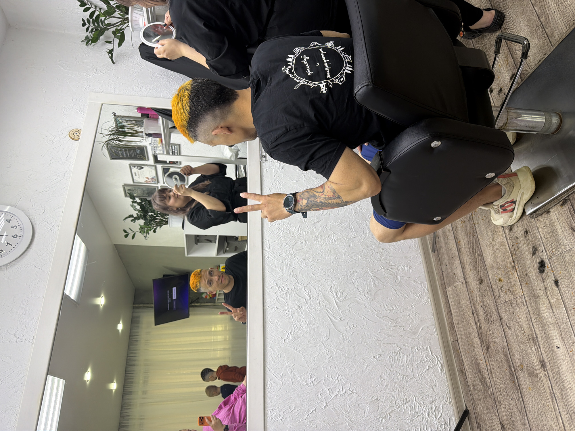 The image shows a man with a black shirt and blonde-tipped hair in what appears to be a barber shop or hair salon. He is sitting in a black salon chair, giving a peace sign. He has tattoos on his left arm and is wearing a watch. A mirror in front of him reflects the scene, including the man and a stylist holding a hand mirror. Other people are faintly visible in the mirror's reflection. The floor is wooden and covered with hair clippings. The wall is textured white. There are plants, certificates, and a television visible in the background via the mirror reflection. The location is Chaikovsky, Russia.