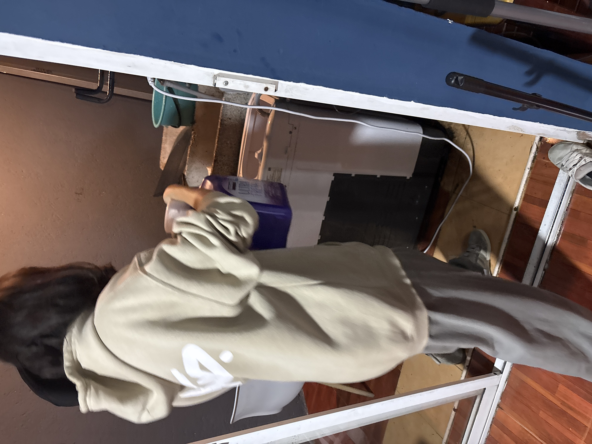 A person wearing a beige hoodie and gray sweatpants stands in an indoor space. The person is viewed from above, with their head towards the left side of the image. Their dark hair is visible under what appears to be a dark-colored cap. A white container with a blue top is visible next to the person. The person's feet, shod in gray sneakers, are visible on a floor with wood paneling. The area is cluttered with various items including an electrical outlet, a white electrical device, a blue surface, and possibly cleaning supplies. The scene is inside an enclosed area. There is no information in the image to identify the location as San Andrés Cholula, Mexico.