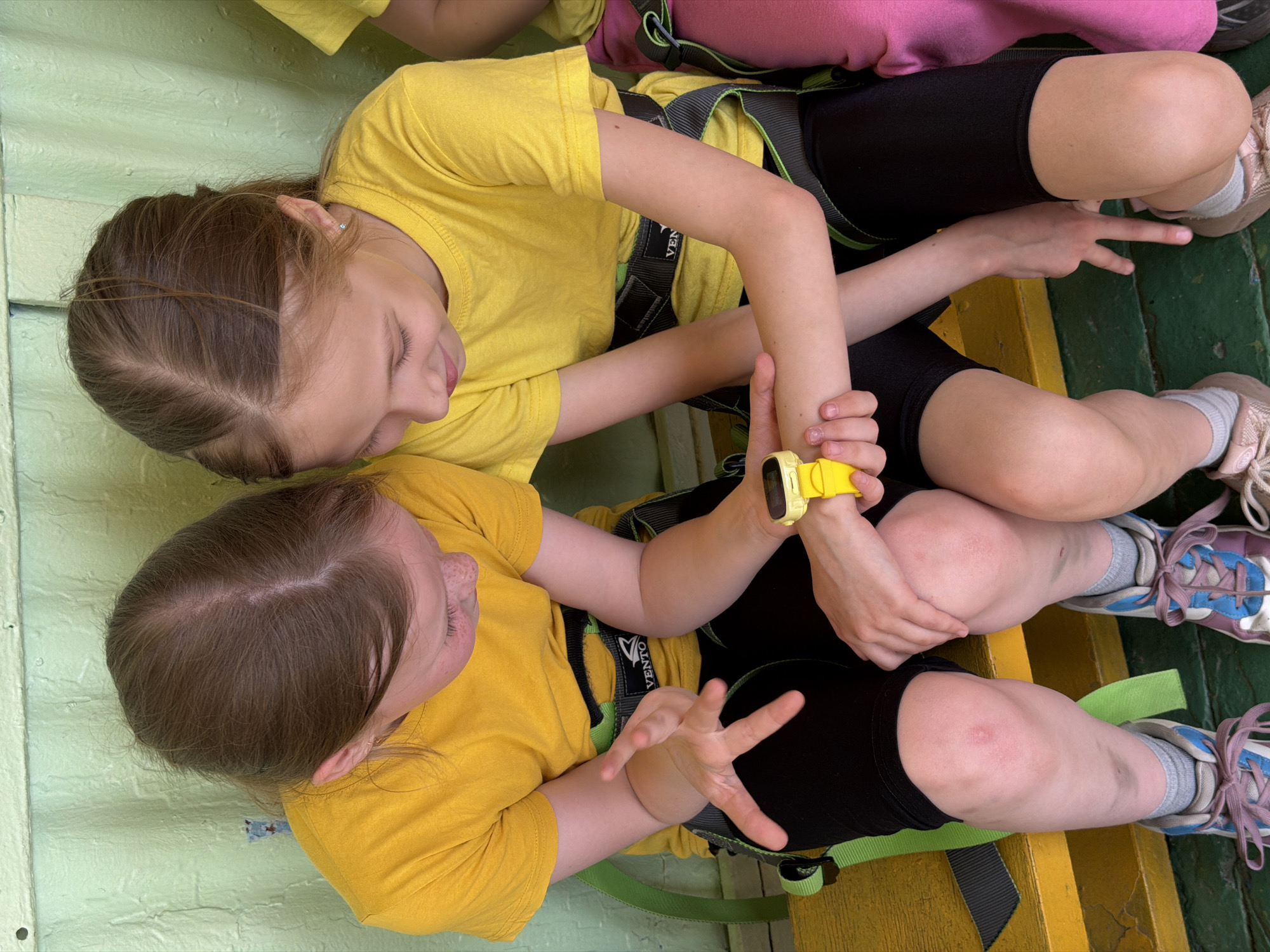 Two young girls, seen from above, are seated side-by-side. Both are wearing yellow t-shirts, black shorts, and climbing harnesses. The girl on the right has brown hair pulled back in a braid, and she is holding the wrist of the other girl, who has a yellow watch on her wrist. The girl on the left is holding up her hand, fingers spread apart. The girls are sitting on a platform, with a green painted surface and yellow accents. Based on the harnesses and platform construction, the location is likely a high ropes course or adventure park, possibly in Izhevsk, Russia, though that cannot be confirmed from the image.