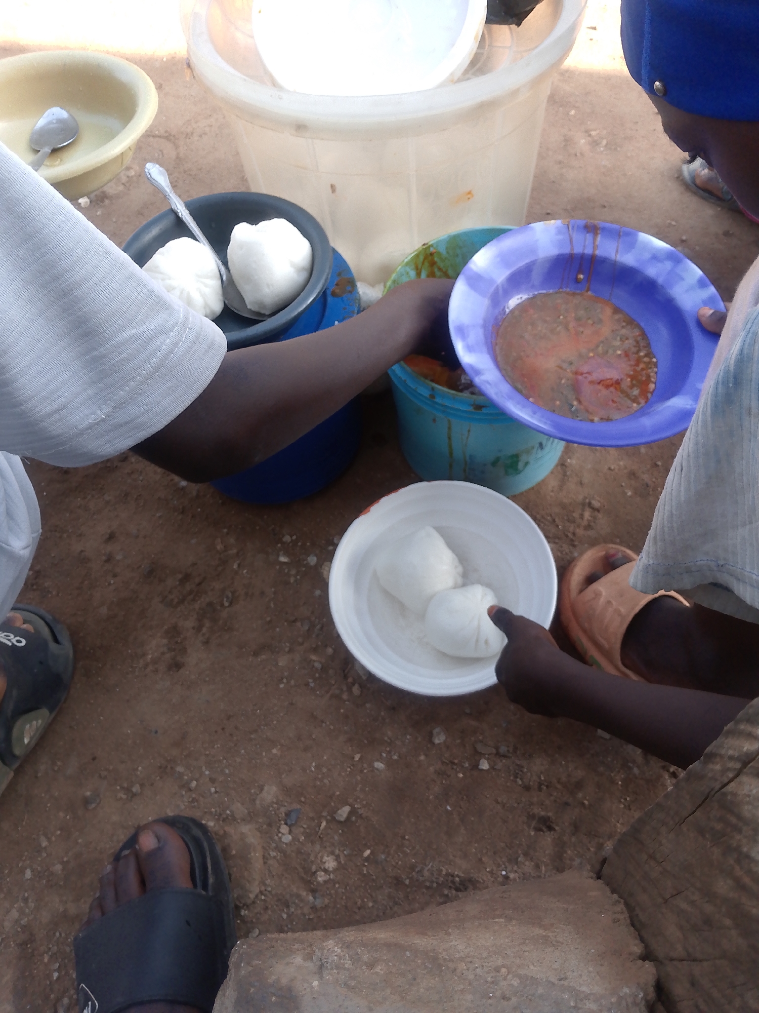 The image captures an outdoor scene, likely in Abuja, Nigeria, depicting a meal being served. The ground appears to be dirt with scattered small stones and debris.
In the foreground, a person's feet are visible. One foot wears a black sandal with a white logo. Another foot, bare, is also in a black sandal.
Several bowls and containers are arranged on the ground. A light yellow bowl with a spoon inside is to the left. Next to it is a dark blue bowl containing two white, rounded food items, possibly fufu or pounded yam, with a silver spoon resting on them. A larger, pale blue bucket-like container holds a reddish-brown stew or sauce. A purple plate, held by a person whose arm is partially visible, contains a similar reddish-brown liquid with bits of ingredients. Another white bowl is placed on the ground, holding three white, rounded food items.
The upper portion of the image shows a person wearing a white garment, possibly a shirt or a smock, with textured fabric. Another person, wearing a blue head covering, is partially visible on the right, seemingly observing the meal.
The overall impression is one of a communal or informal meal preparation and serving setting in an outdoor environment.