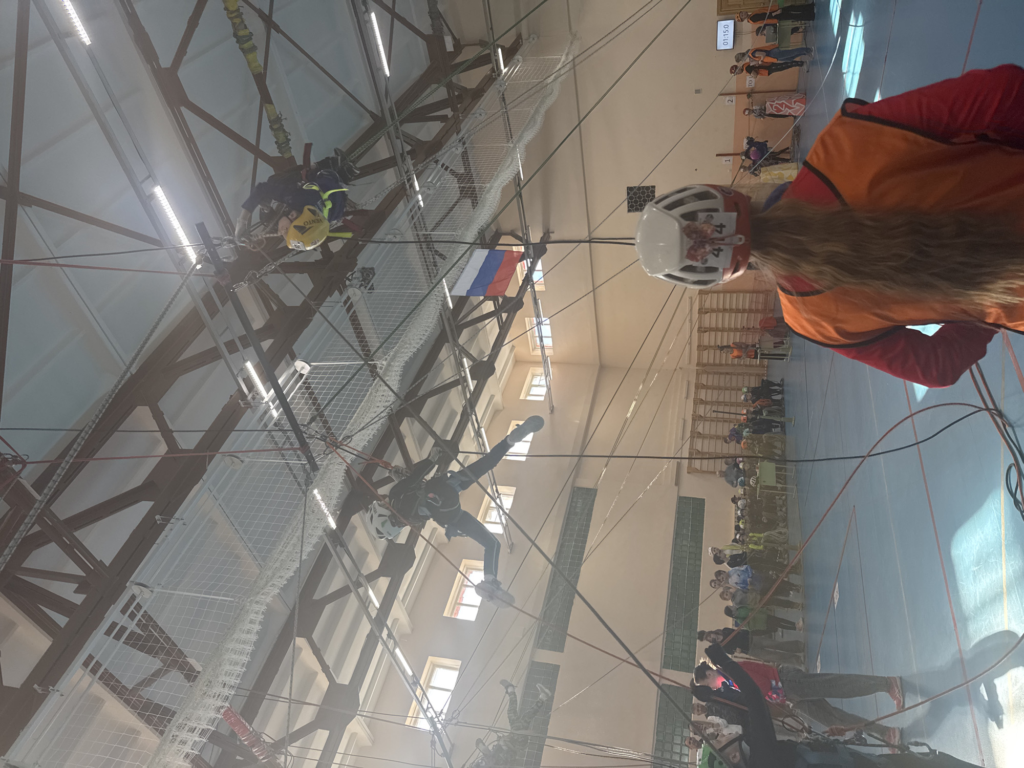 The image shows an indoor sports facility in Usolye, Russia, possibly a gymnasium. It appears to be a competition or demonstration involving rope climbing and related activities.
Several individuals are suspended in the air via rope systems attached to the ceiling structure. One person in the foreground wears a helmet with the number "44" visible, an orange vest, and has long hair. They seem to be a participant or staff member.
In the background, there's a group of spectators observing the activity. The Russian flag is visible, hanging near the upper structure. The walls are light-colored, and the ceiling is high, supported by a network of beams and ropes. The floor appears to be a blue gymnasium floor. A digital display screen can be seen, likely showing the timing or scores of the event.