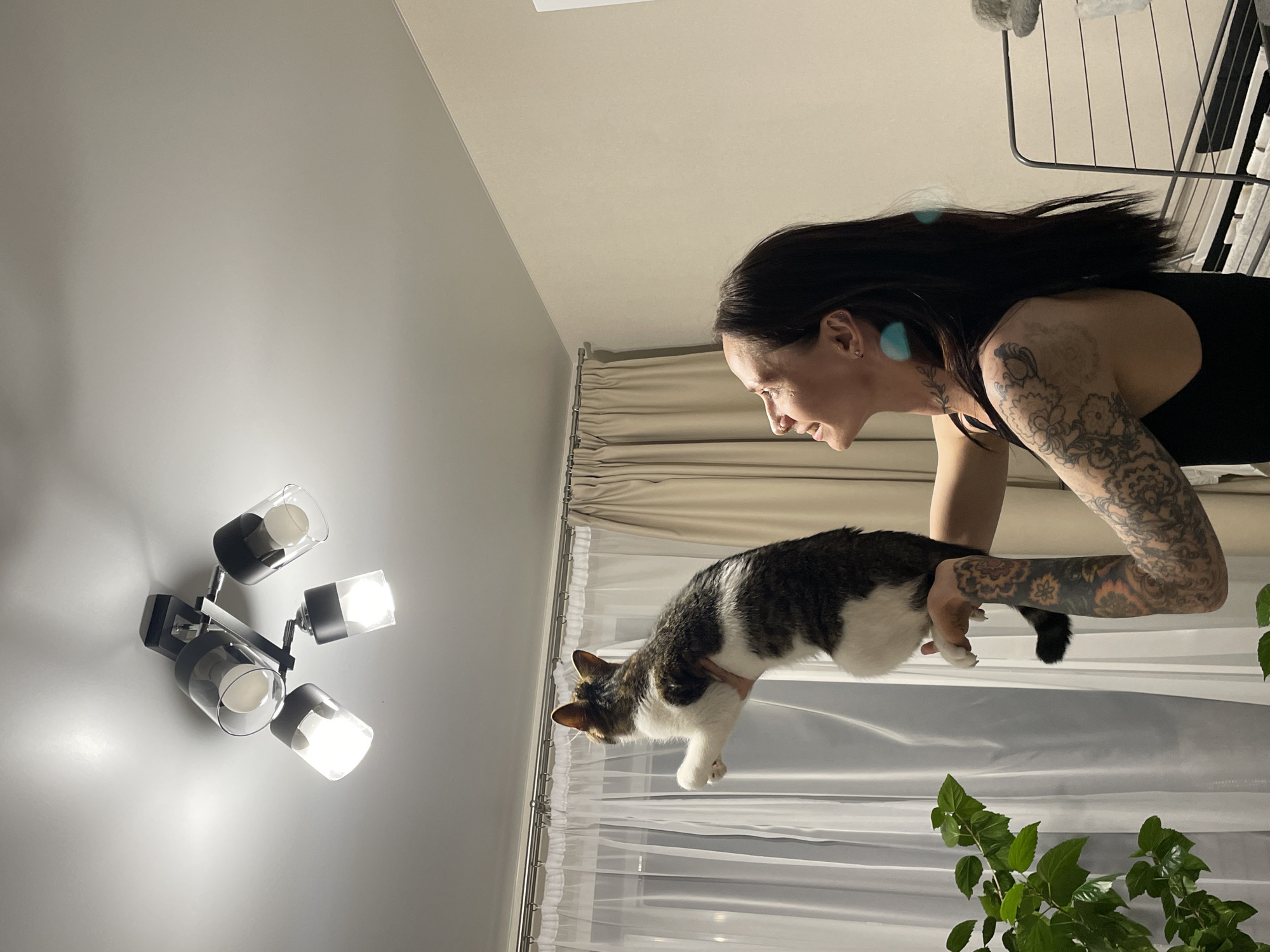 A woman with dark hair and tattoos on her arms is holding a cat up towards a ceiling light fixture. The room has light-colored walls, curtains, and a plant. The location is not specified as the city of Pervomaysky, Russia, therefore the location is unknown.