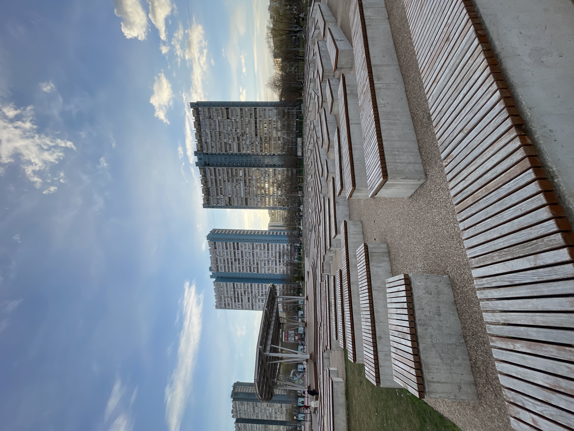 The image shows an outdoor public space in Moscow, Russia. Several Brutalist apartment buildings are visible in the background. A public seating area features rows of concrete benches with wooden slats. The ground is covered in gravel and patches of grass. A few people are visible under a metal structure, possibly a covered stage or seating area. The sky is blue with scattered clouds.
