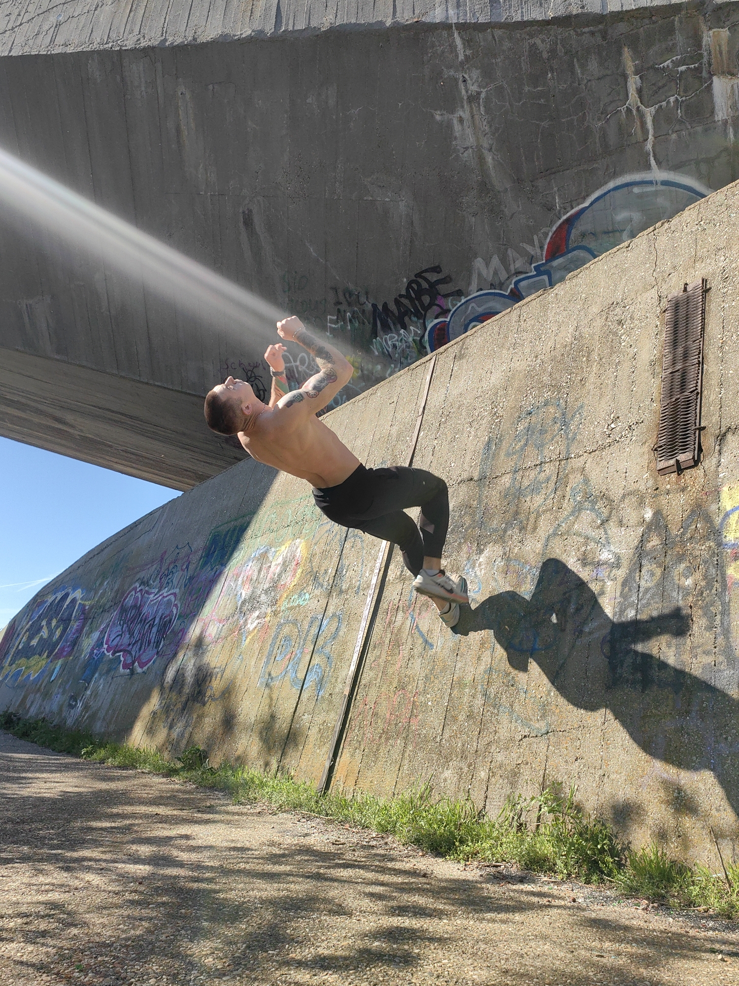 A shirtless man with tattoos on his arms is performing a backflip against a concrete wall covered in graffiti. He is wearing black athletic pants and gray sneakers. His shadow is cast on the wall to his right. Above him is a concrete bridge or overpass. The setting appears to be outdoors on a gravel or dirt path, with grass growing along the base of the wall. Sunlight streams in from the upper left of the frame. The city location in Varna, Bulgaria, cannot be determined from the image.