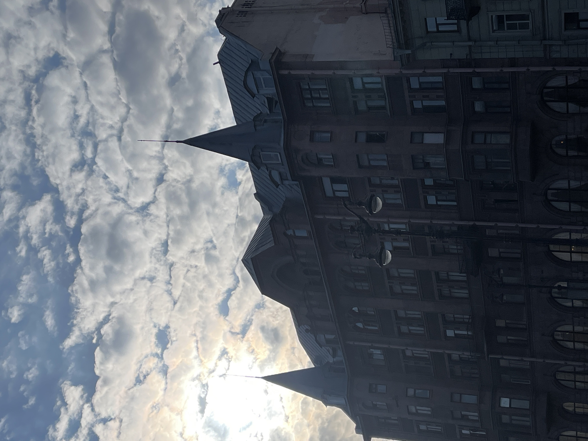 The image shows a building with pointed turrets and a multitude of windows in St. Petersburg, Russia. The building is dark and imposing against a backdrop of a blue sky filled with scattered white clouds and a bright sun shining through. A street lamp is visible in the foreground.