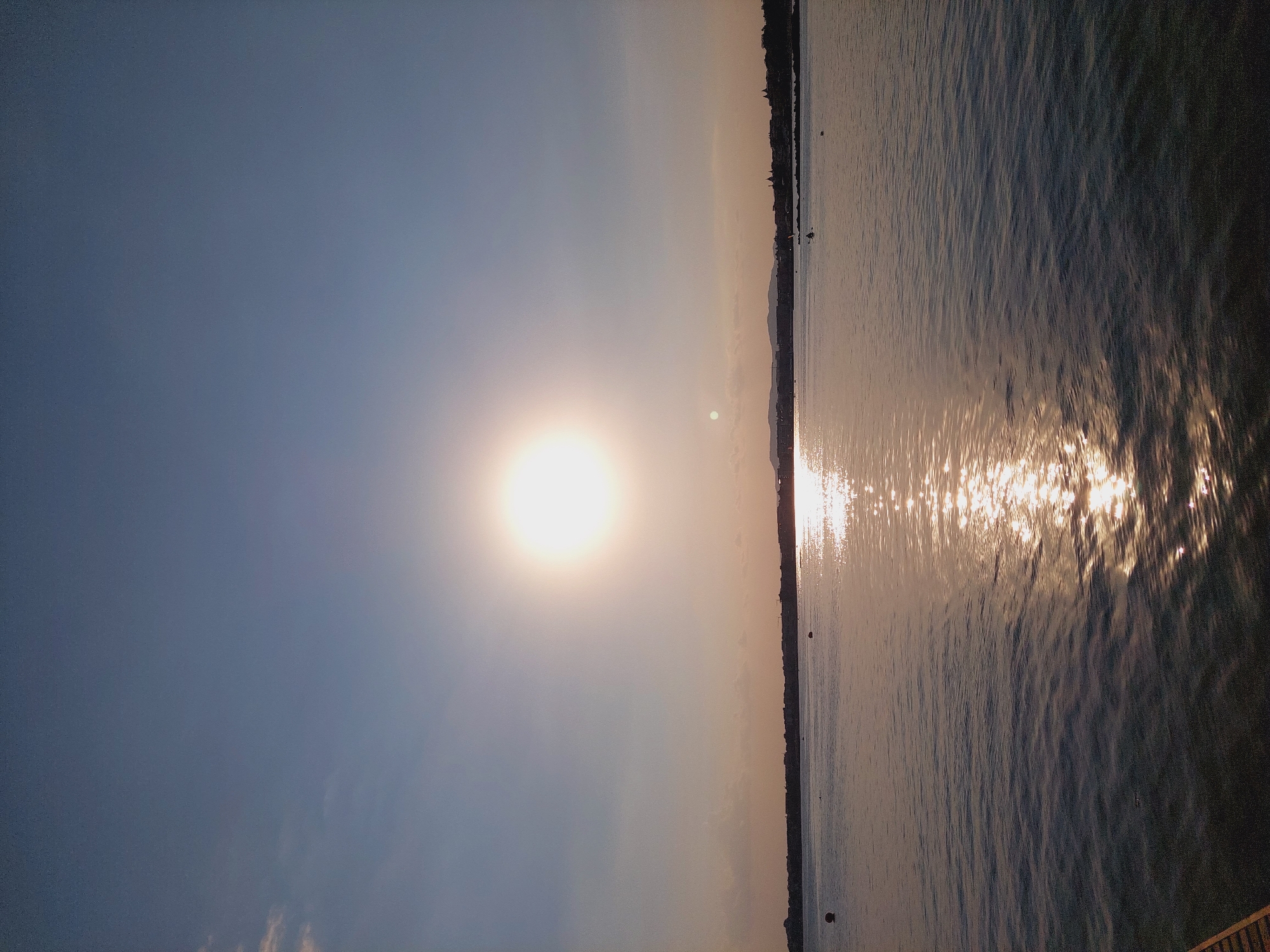 A wide-angle vertical shot captures a tranquil sunset over the Black Sea, viewed from Nesebar, Bulgaria. The sun, a bright, luminous orb, sits high in the pale blue sky, casting a shimmering golden path across the gently rippling water. A faint haze softens the horizon, where a distant, silhouetted coastline with buildings and a crane is visible. The water itself is a deep, reflective blue, with subtle waves creating a textured surface. In the foreground, the edge of a wooden structure, possibly a pier or boardwalk, is visible on the lower left. A few dark buoys dot the water, marking an area of calm. The overall atmosphere is serene and peaceful, with the fading light creating a warm and inviting scene.