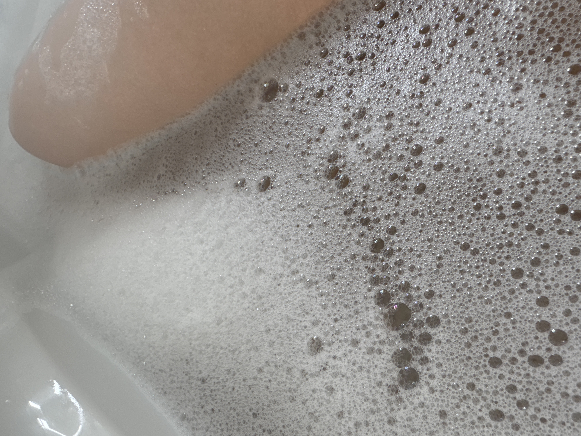 The image shows a close-up view of a bathtub filled with soapy water and bubbles. A portion of a person's leg is visible in the upper left corner. The bubbles vary in size, creating a textured surface. The scene is indoors, presumably in a bathroom. There is no indication of the location being in Desborough, United Kingdom.