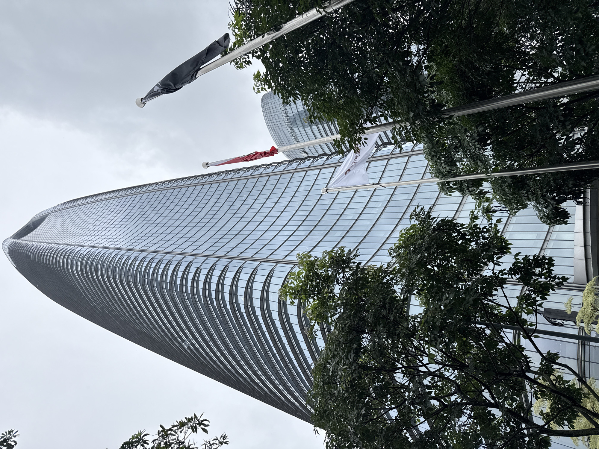 This is a low-angle shot of a modern, curved skyscraper in Haizhu District, China. The building's facade is made of reflective glass panels arranged in a grid pattern, creating a sleek and futuristic appearance. The bottom of the skyscraper curves inward, giving it a unique, organic shape. Several flags are visible flying on poles in front of the building, including a black flag, a red and white flag, and a white flag with some text that is not legible. Lush green trees with dense foliage are in the foreground, partially obscuring the view of the building and adding a natural element to the urban scene. The sky is overcast and appears a muted gray, suggesting cloudy or perhaps rainy weather. The lighting is soft and diffused, typical of an overcast day. The image captures a view looking upwards, emphasizing the scale and architectural design of the skyscraper against the sky. There are no people visible in the frame, and the focus is on the architecture and the surrounding natural elements. The overall mood is one of modern architectural ambition blended with nature.