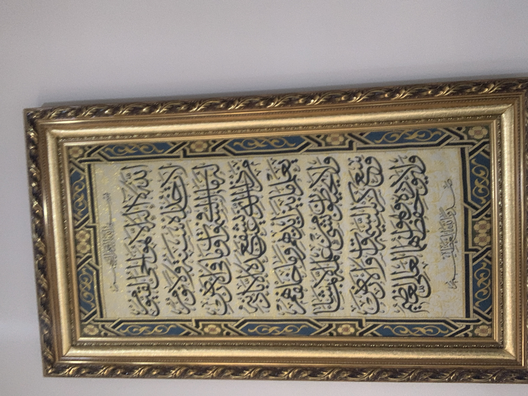 This is a framed piece of Islamic calligraphy, likely a page from the Quran or a religious text. The calligraphy is in black ink on a light-colored background, with decorative borders. The frame is ornate and golden, with intricate patterns. The calligraphy is arranged in multiple columns and appears to be quite dense. The overall impression is one of religious devotion and artistic craftsmanship. The piece is hanging on a plain, light-colored wall, suggesting it is displayed indoors as a decorative or devotional object. There is no indication of the time of day, weather, or specific location beyond the context of being in Abuja, Nigeria. The scene is static and the mood is likely reverent or peaceful, given the subject matter. No people are visible in the image.
