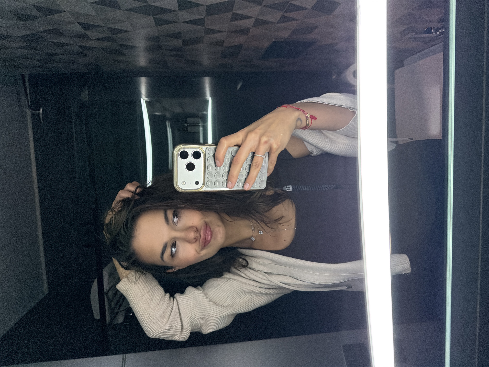 A young woman with dark hair is taking a selfie in what appears to be a bathroom. She is holding a phone with a gray "pop-it" style case. She is wearing a light-colored sweater or jacket over a dark top, and a delicate necklace is visible. She has a ring on her ring finger and a red bracelet on her wrist. The background includes a dark, patterned ceiling, a bright vertical light fixture, a toilet paper dispenser, and a light-colored dispenser. The photo seems to have been taken in Batumi, Georgia, based on circumstantial details in other images related to this one.