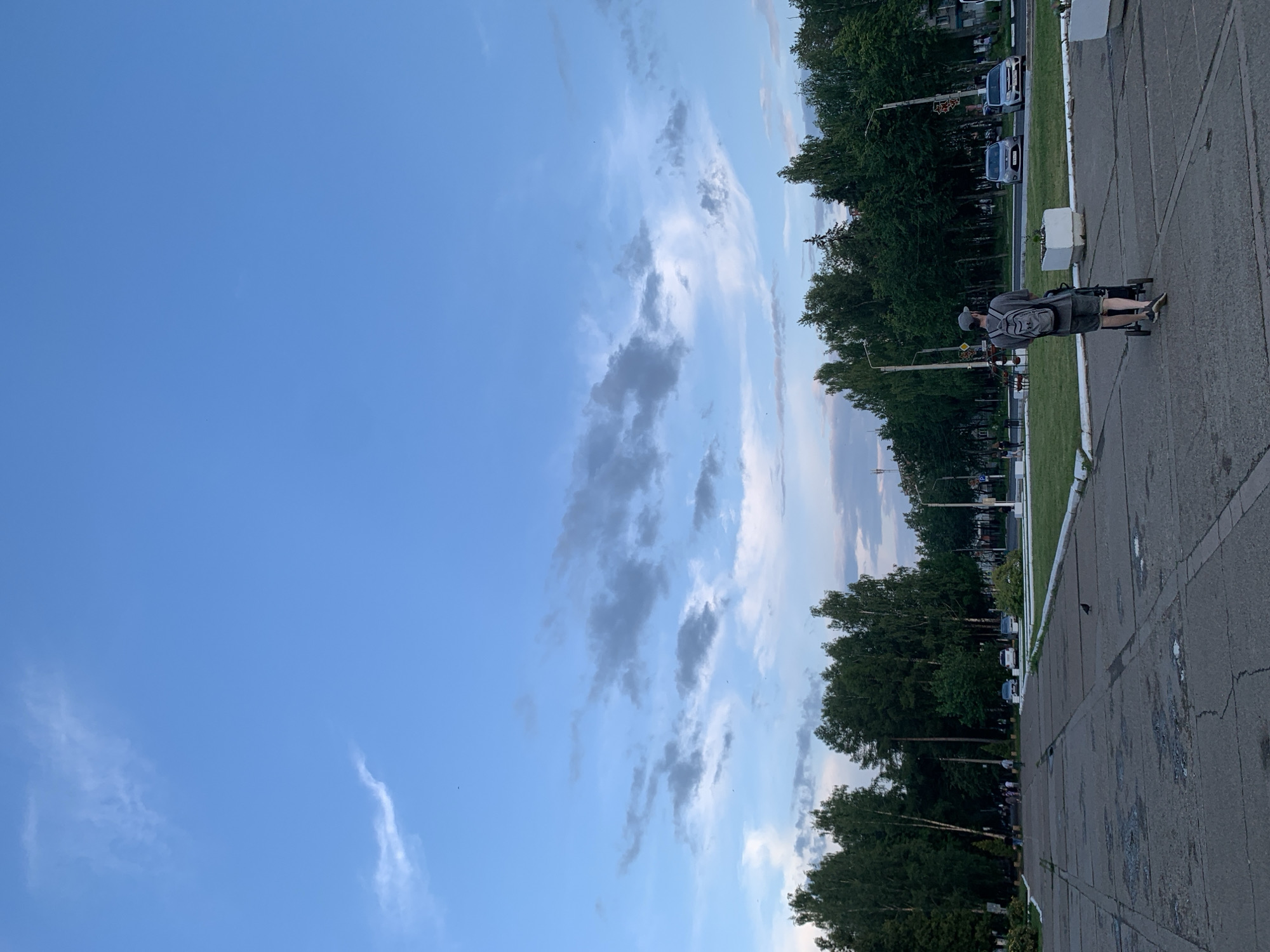 An outdoor, eye-level, medium shot captures a person from behind, pushing a stroller on a paved area in Chaikovsky, Russia. The individual is wearing a grey t-shirt, grey shorts, a grey baseball cap, and a grey backpack. They are walking away from the camera towards a street lined with trees. The paved area appears to be a public square or walkway, with cracks and patches visible on the concrete. A pigeon is on the ground in the foreground. In the background, there is a row of green trees, a street with cars, and buildings. The sky is blue with scattered clouds. The image suggests a late afternoon or early evening setting due to the lighting.