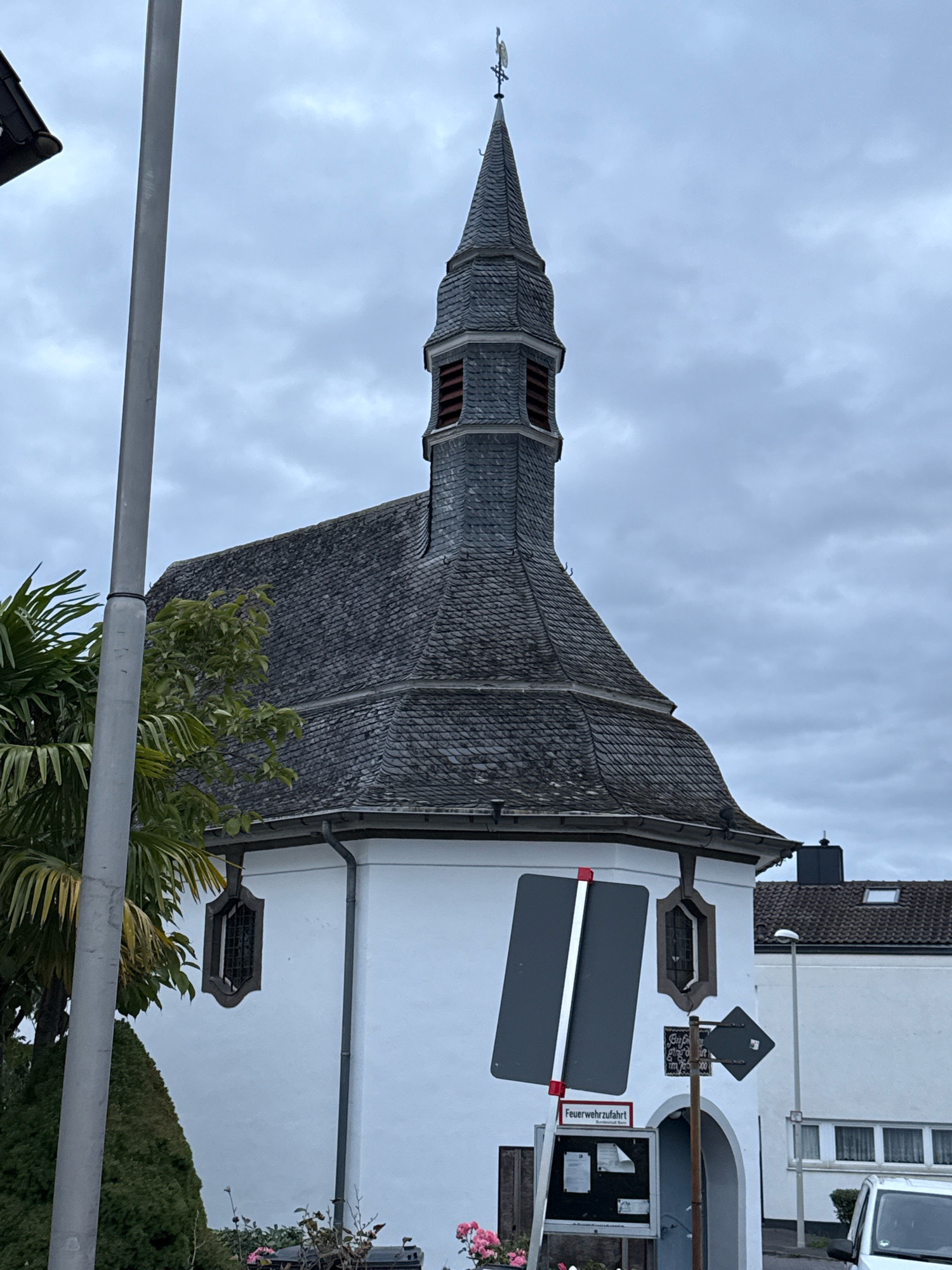 The image shows a building with a distinctive steeple, likely a church or chapel, in Bonn, Germany. The building has a white facade and a dark, slate-tiled roof. The steeple is tall and pointed, with a weather vane at its peak. The sky is overcast, with dark clouds indicating a potentially rainy day. In the foreground, there is a street lamp and some greenery, including palm trees and other bushes. There are also some signs visible, one of which says "Feuerwehrzufahrt" which means "fire department access" in German. The scene is quiet and appears to be an exterior shot of a public building.