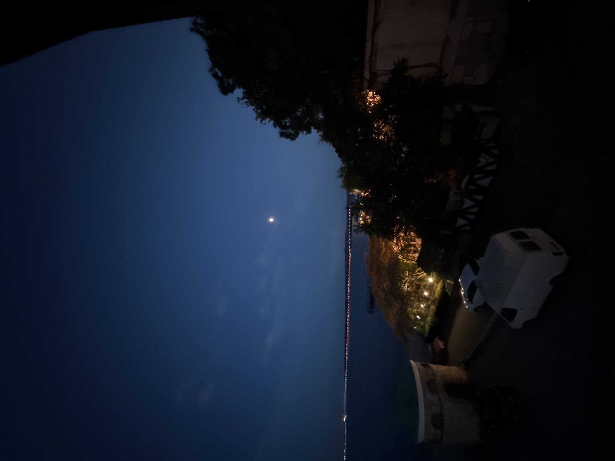 The image depicts a nighttime scene overlooking a body of water, possibly the Kerch Strait, with a dimly lit coastline featuring a string of lights. A crescent moon is visible in the deep blue sky, along with some faint clouds. In the foreground, a paved area shows several vehicles, including a white van and a white car. To the left, there is a round, stone structure with a turquoise roof, resembling a small pavilion or turret. Trees with illuminated foliage are situated behind the paved area and vehicles, suggesting an outdoor dining or entertainment space. A dark, dense tree canopy occupies the right side of the frame, partially obscuring a building with a light-colored facade. The overall impression is of a serene, nocturnal landscape near the coast.