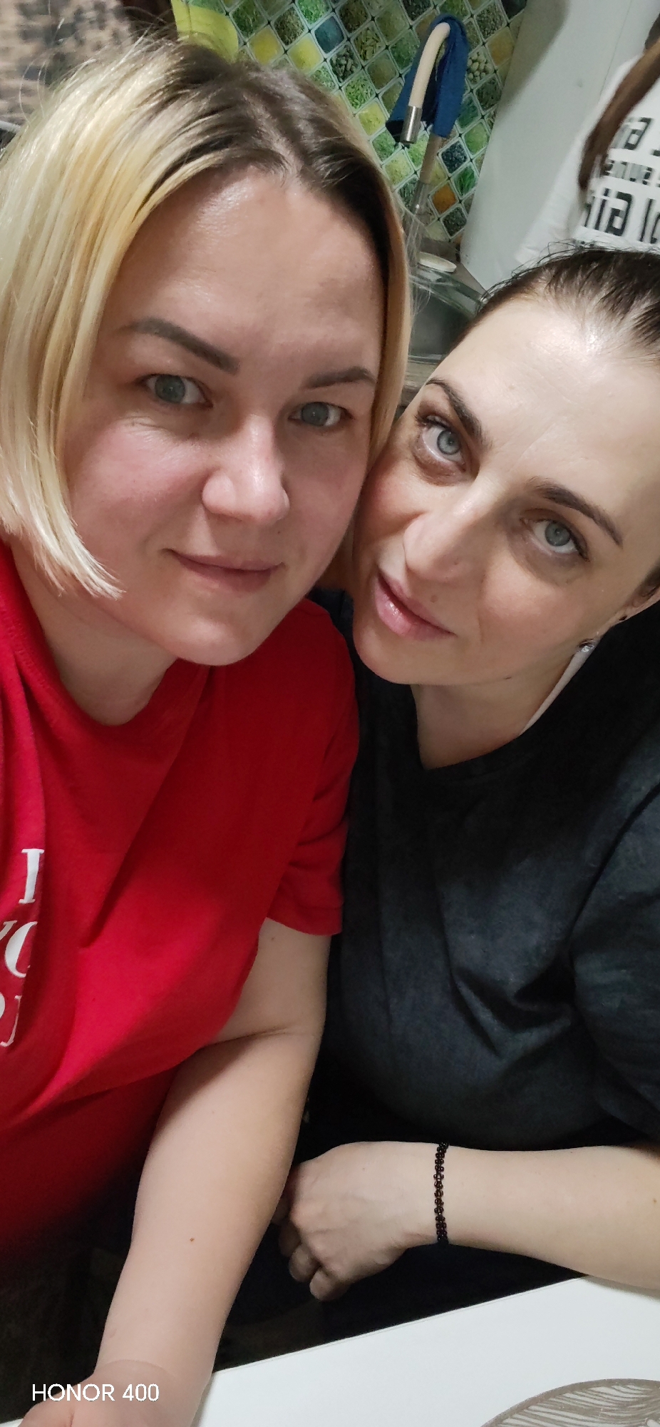This image, captured in Noviy, Russia, presents two adult females in a close-up, selfie-style photograph within an indoor setting, likely a kitchen or utility area. The female positioned on the left has blonde, shoulder-length hair with visible dark roots, light-colored eyes, and a fair complexion. She is wearing a red short-sleeved t-shirt with partially visible white text on the front and displays a slight smile. The female on the right has dark hair pulled back, light-colored eyes with visible eye makeup, and a fair complexion. She is wearing a dark grey short-sleeved t-shirt over a white undershirt and a dark, possibly beaded, bracelet on her left wrist. She also exhibits a slight smile and is positioned closely to the left female. Both individuals are looking directly at the camera. The background features a tiled wall with a mosaic pattern of green, yellow, and white squares, a white flexible hose, and a partially visible white surface with dark, illegible text. A white countertop or table is visible in the foreground.