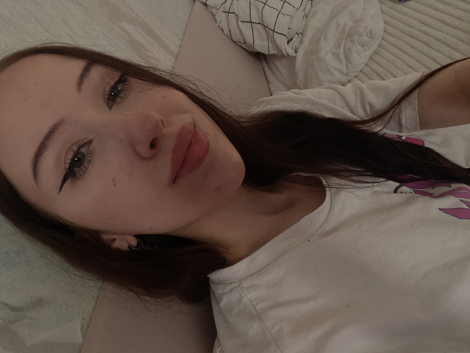 A young woman with long, dark hair lies on a bed, looking towards the camera. Her eyes are open, and she is wearing makeup, including winged eyeliner and mascara. Her lips are slightly parted, and she has a light-colored t-shirt on with a graphic design visible on the right side. The background shows bedding, including a pillow and a comforter with a grid pattern. The scene appears to be indoors, possibly in a bedroom. The lighting suggests it might be evening or a dimly lit room. The woman's expression is relaxed, with a slight smile. There is no visible text in the image. The location context is Votkinsk, Russia.